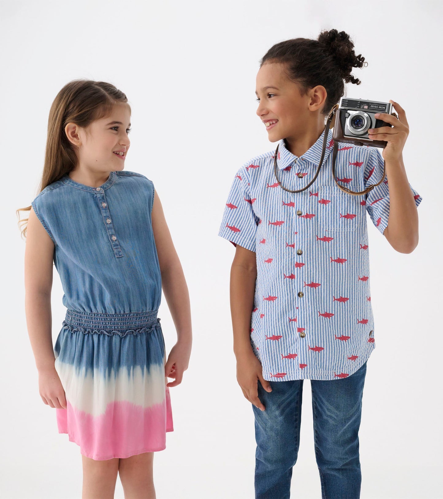 Girls' sleeveless denim dress with smocked waist and pink ombre hem, paired with a boy in a striped shirt and jeans.