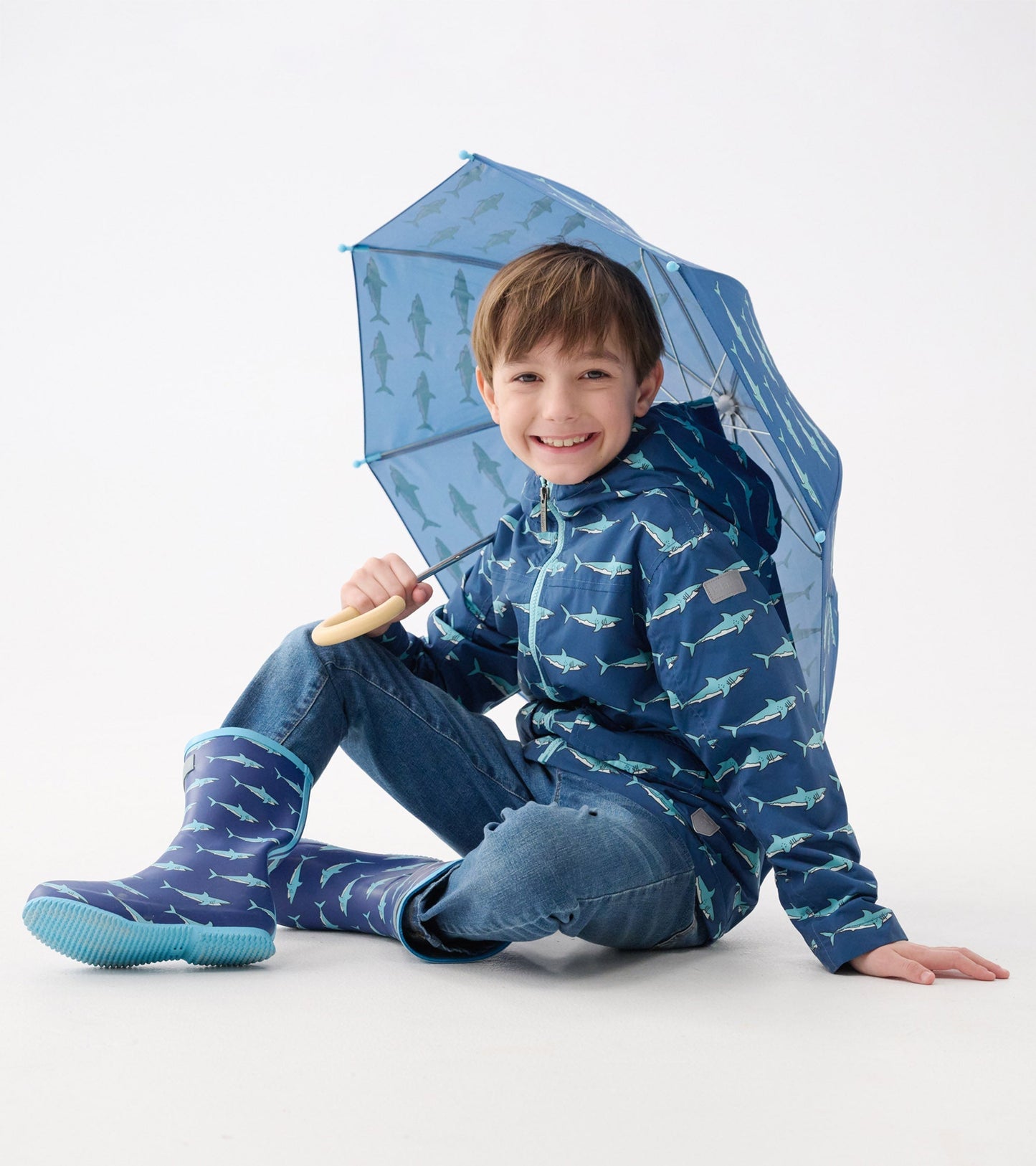 Lightweight blue raincoat with shark pattern, paired with matching rain boots, worn by a smiling boy holding an umbrella.