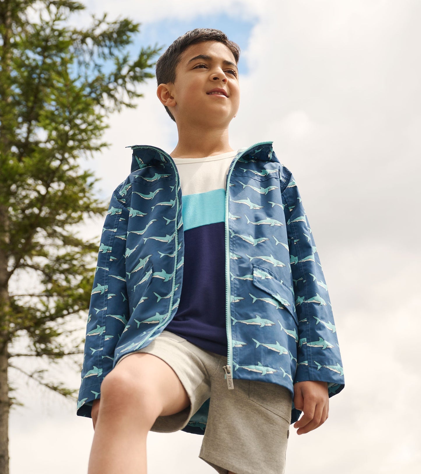 Lightweight blue raincoat with shark pattern, zip-up front, worn by a boy outdoors, paired with a t-shirt and shorts.