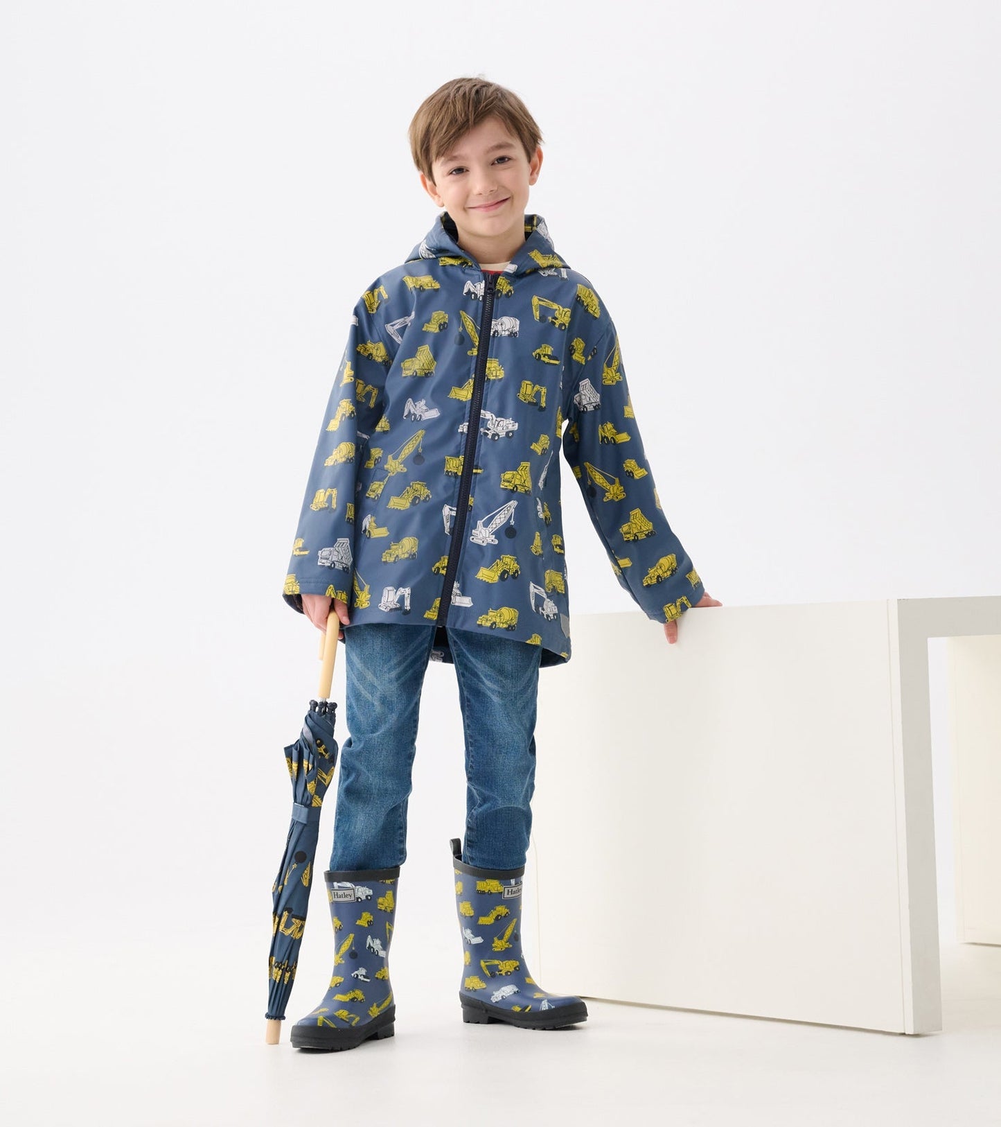 Blue zip-up raincoat with yellow construction vehicle patterns, paired with blue rain boots and holding an umbrella.