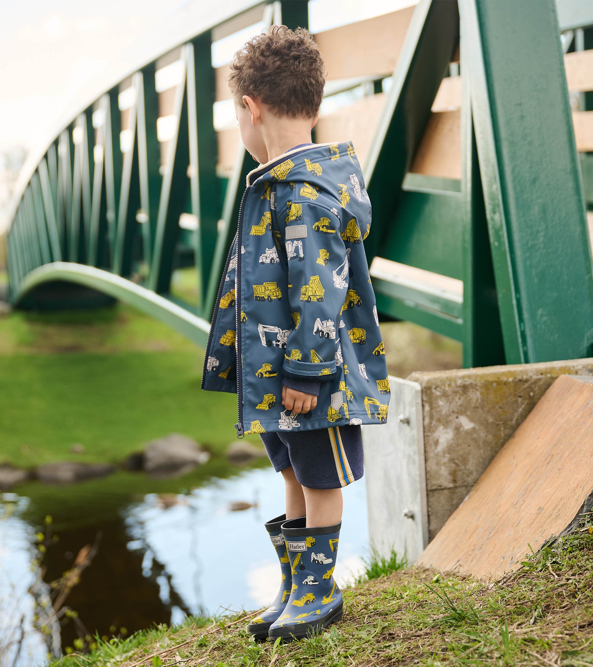 Blue zip-up raincoat with yellow construction vehicle patterns, worn by a child standing by a pond.