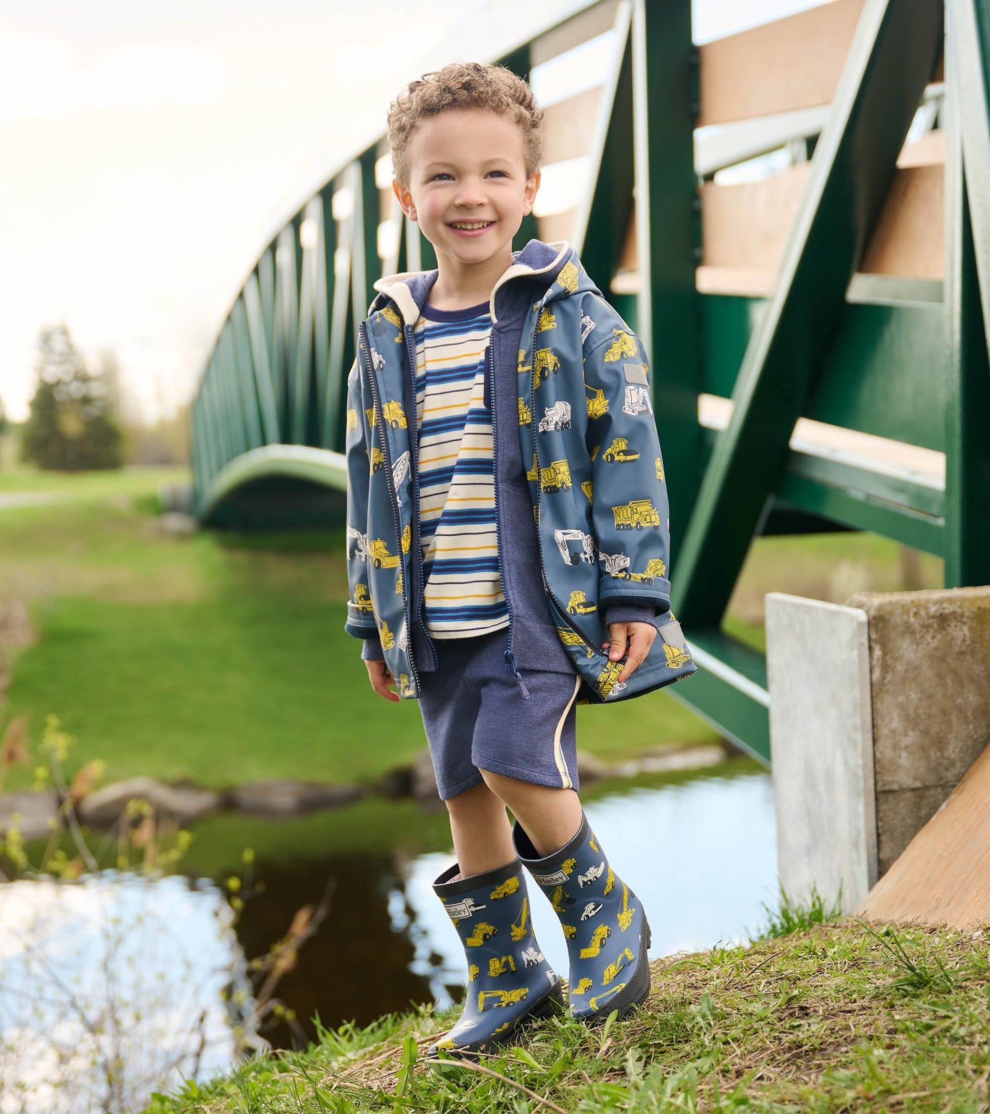 Blue zip-up raincoat with construction vehicle print, worn over a striped shirt and shorts, paired with matching rain boots.