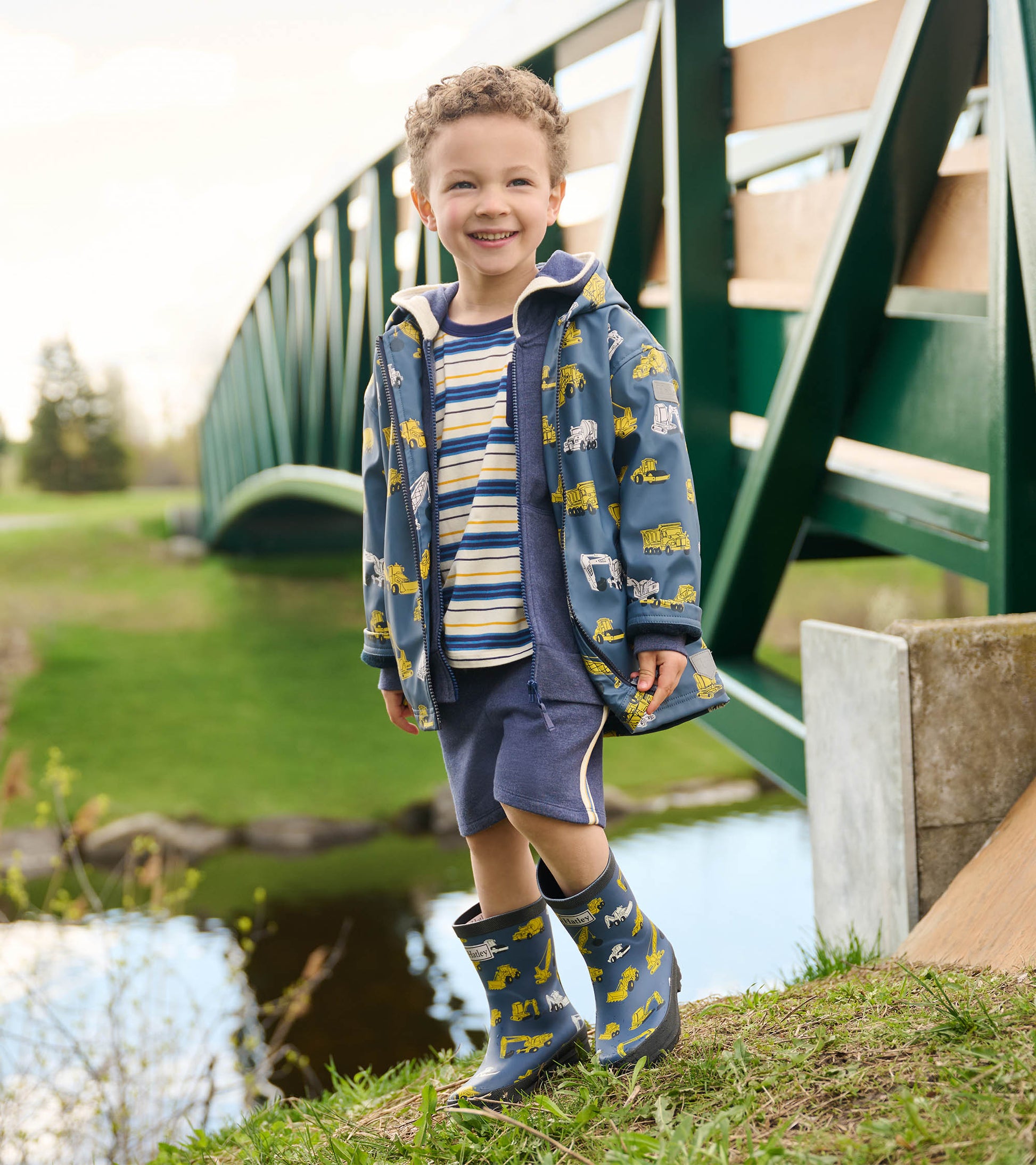 Blue zip-up raincoat with construction vehicle print, worn over a striped shirt and shorts, paired with matching rain boots.