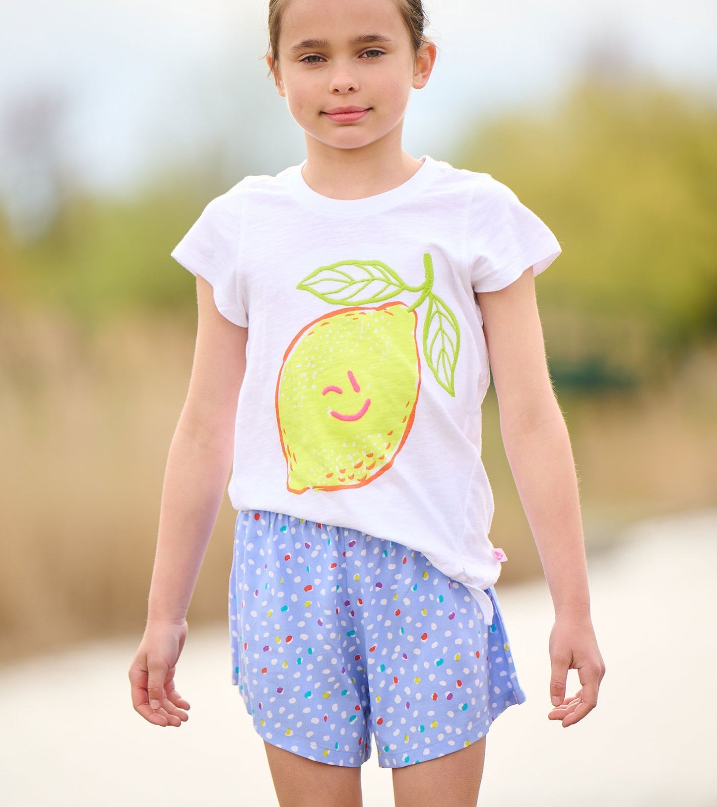 White short-sleeve t-shirt featuring a smiling lemon graphic, paired with blue polka dot shorts.