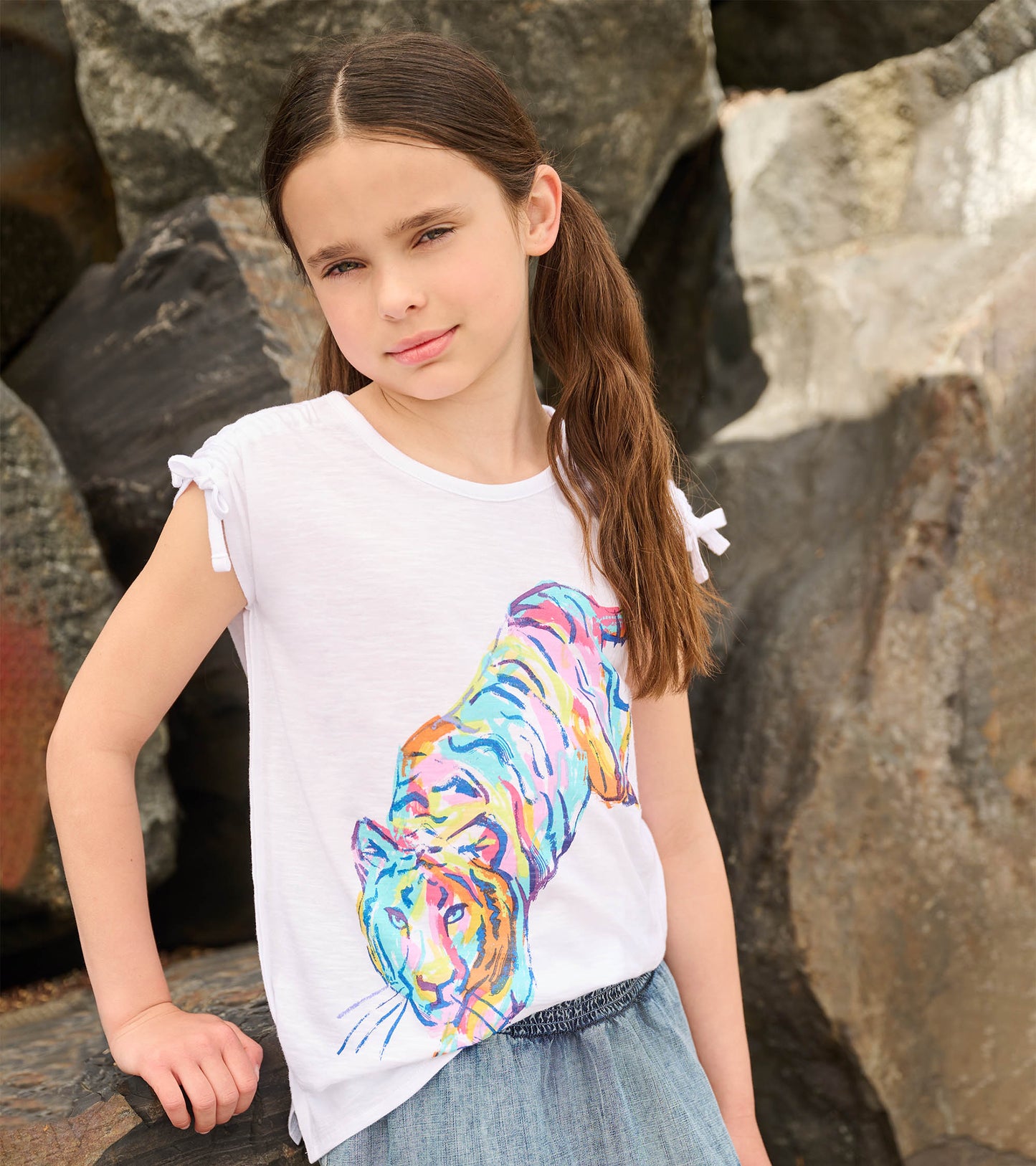 White short-sleeve top featuring a colorful tiger graphic and cinched shoulder details, styled with a denim skirt.