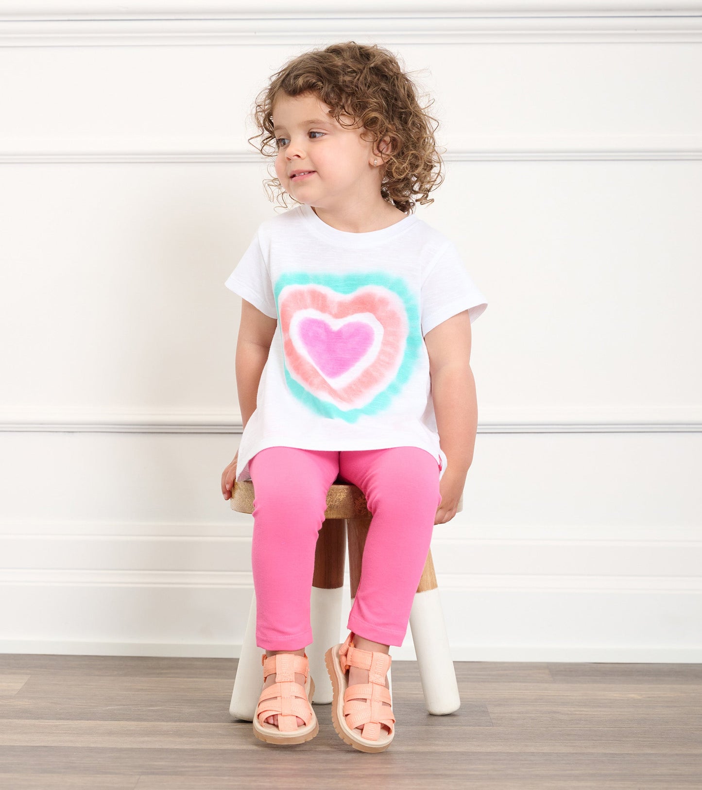 White graphic tee with a colorful heart design, paired with pink leggings and sandals, worn by a toddler sitting on a stool.