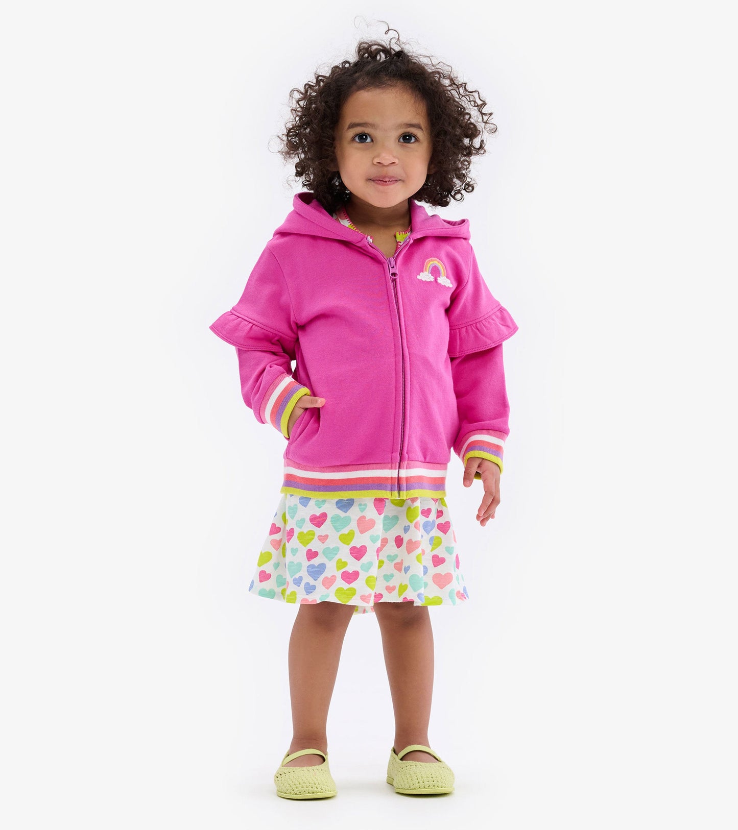 Pink hoodie with flutter sleeves, rainbow detail, paired with a heart-patterned skirt and green shoes.