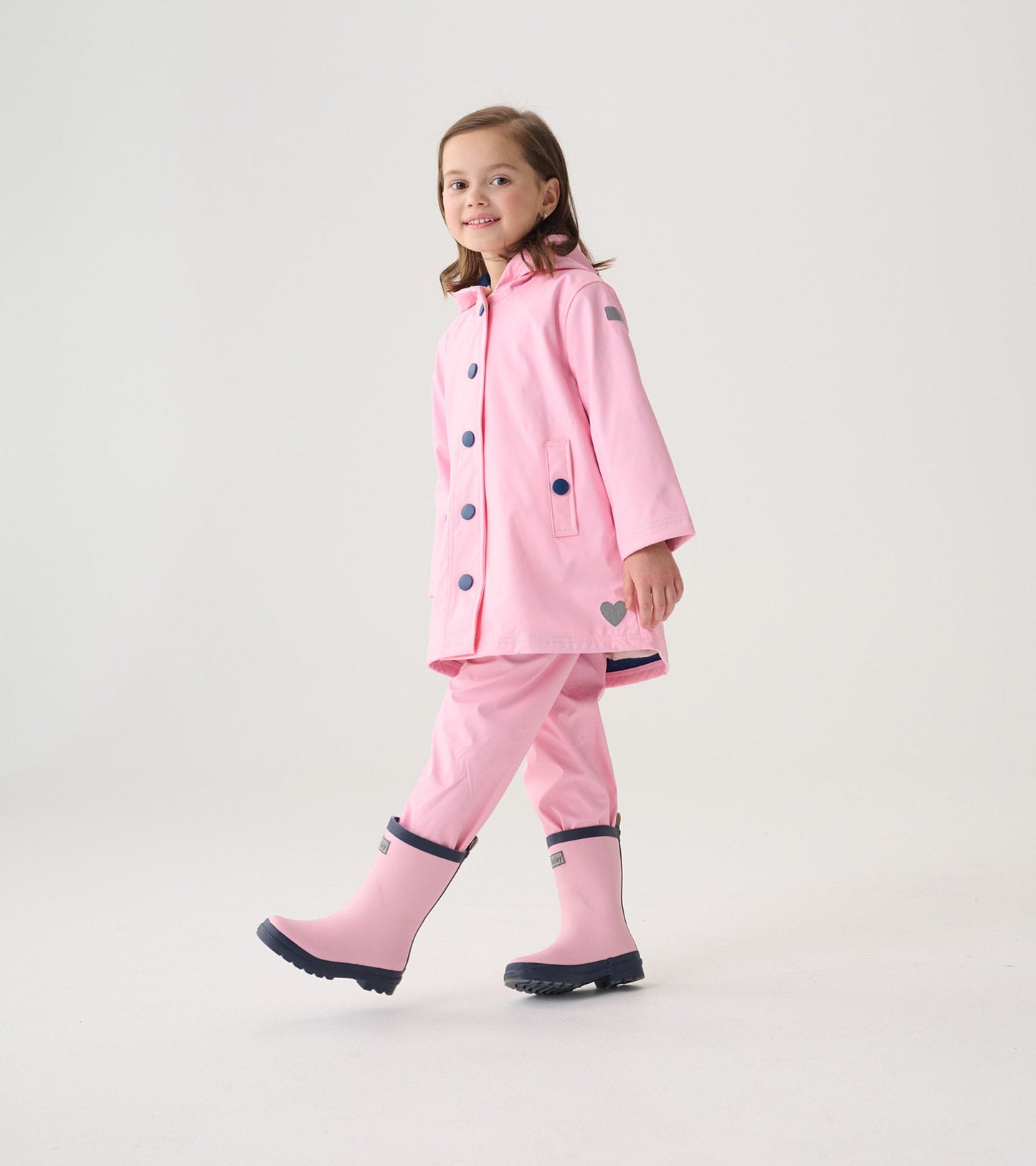 Pink button-up raincoat with navy buttons, paired with matching pink rain pants and boots, worn by a smiling girl.