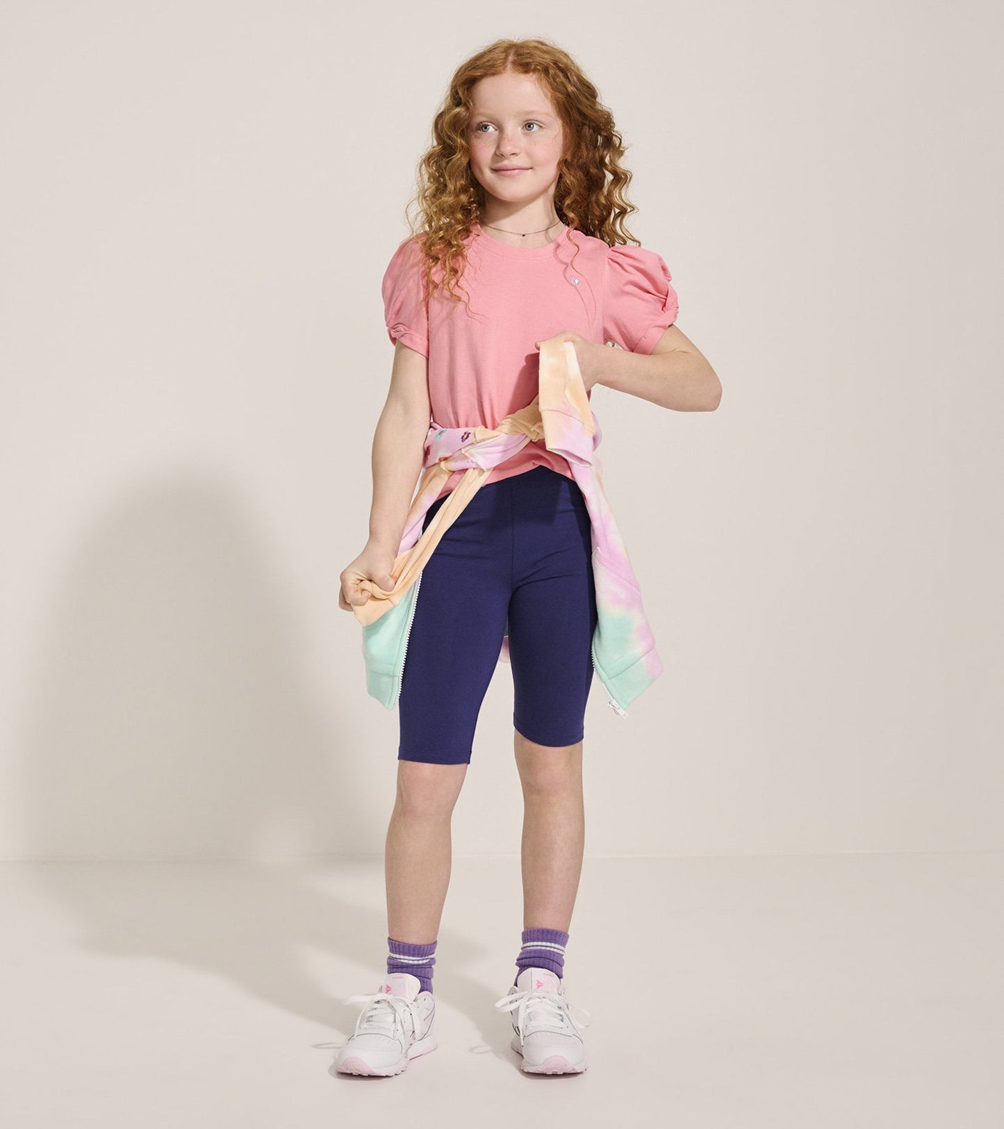 Pink short-sleeve tee with twisted sleeves, paired with navy bike shorts and a pastel tie-dye jacket tied around the waist.