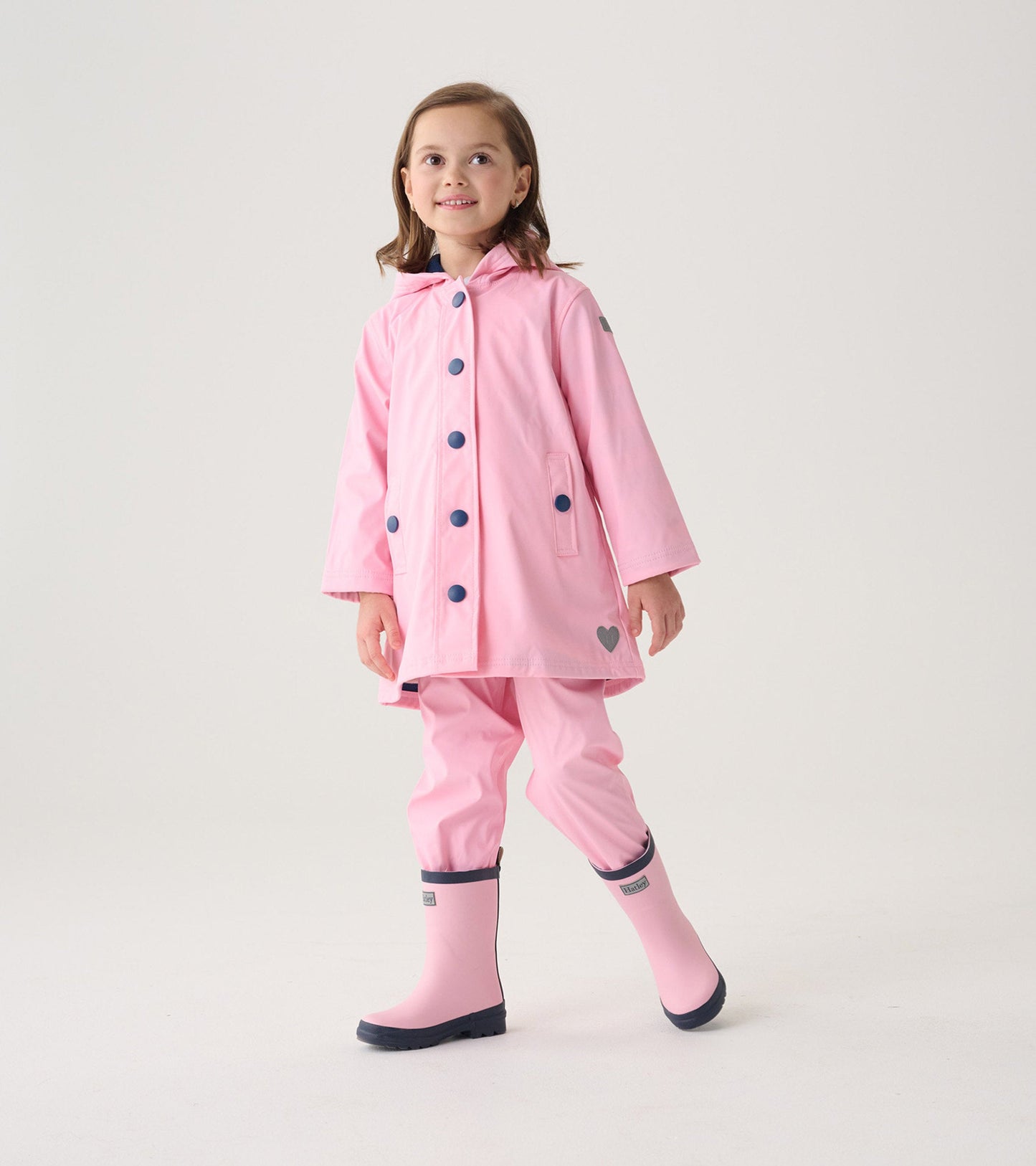 Pink button-up raincoat with navy buttons, hood, and heart detail, worn by a girl with matching pink rain boots.