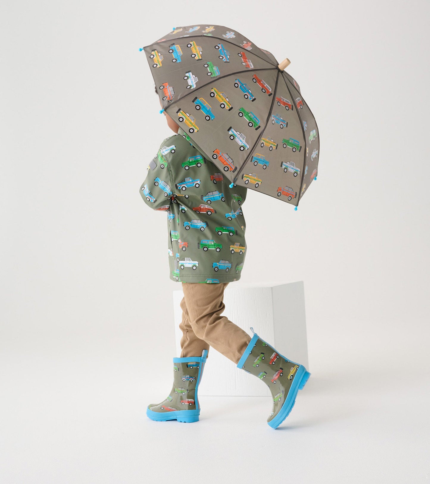 Green wellies with colorful vehicle patterns, paired with a matching umbrella and tan pants, worn by a child.
