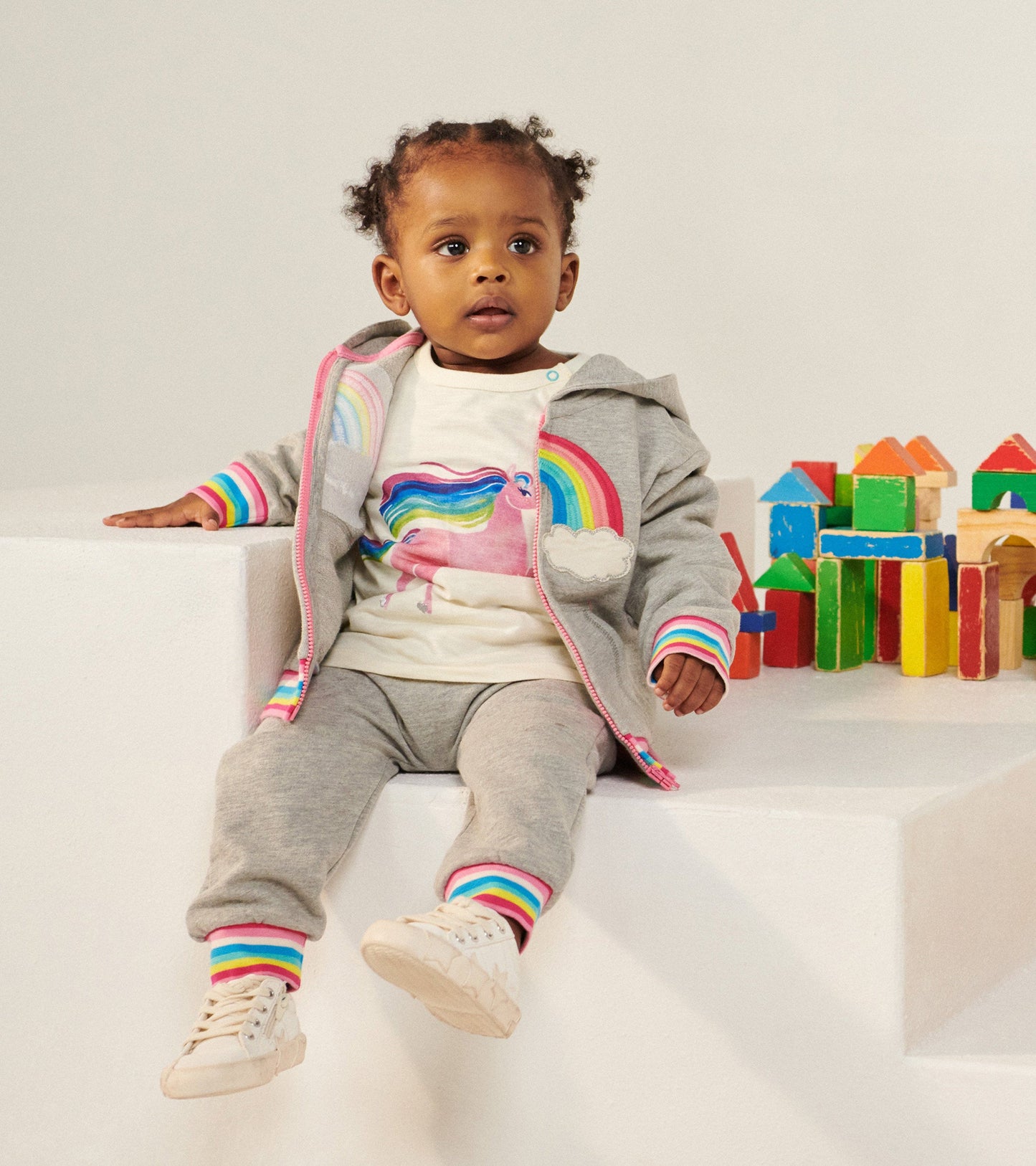 Gray full zip hoodie with rainbow accents, worn by a child sitting beside colorful building blocks.