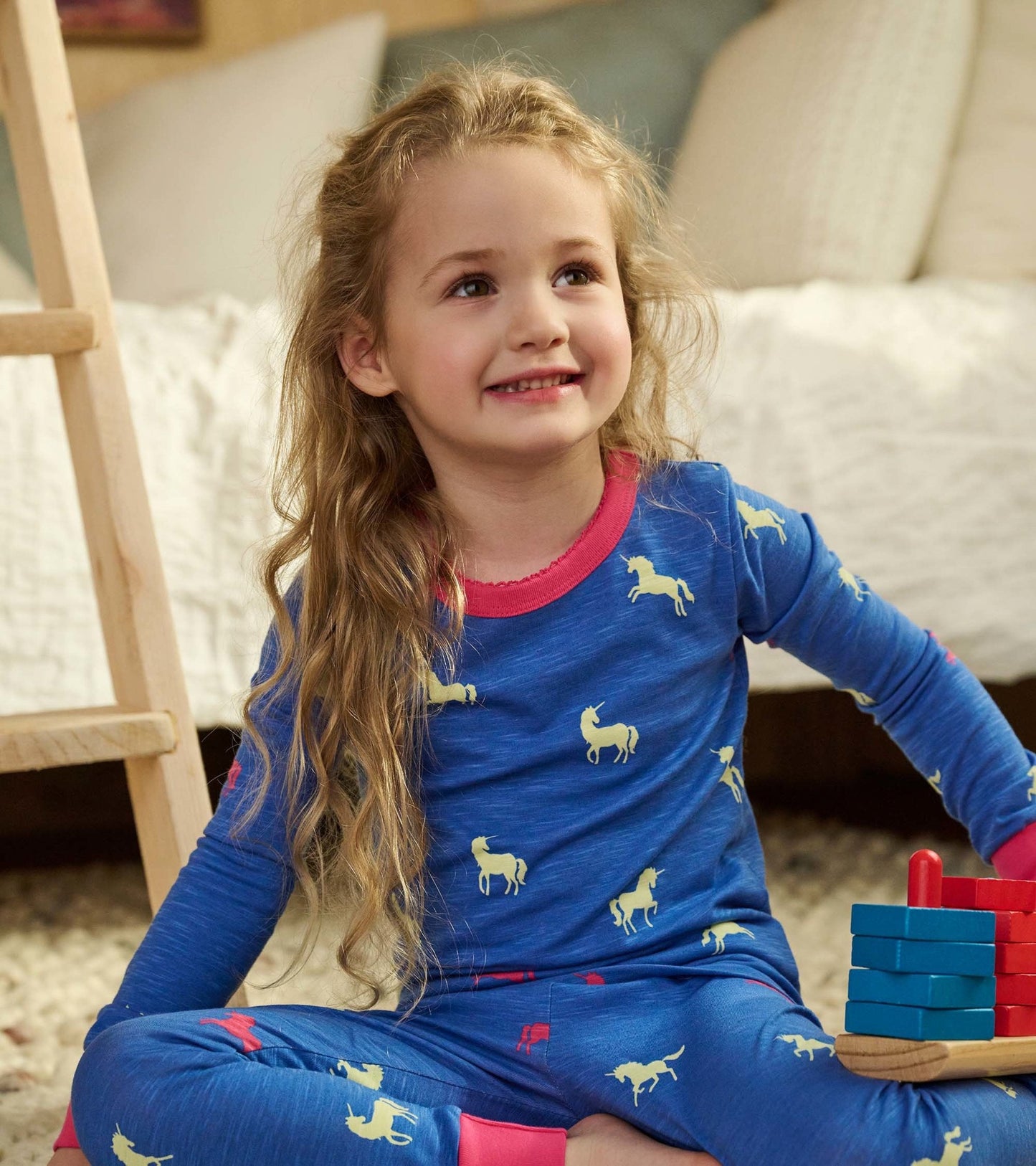 Blue pajama set featuring yellow unicorns, long sleeves, and pink trim, worn by a girl sitting on a rug with toys.