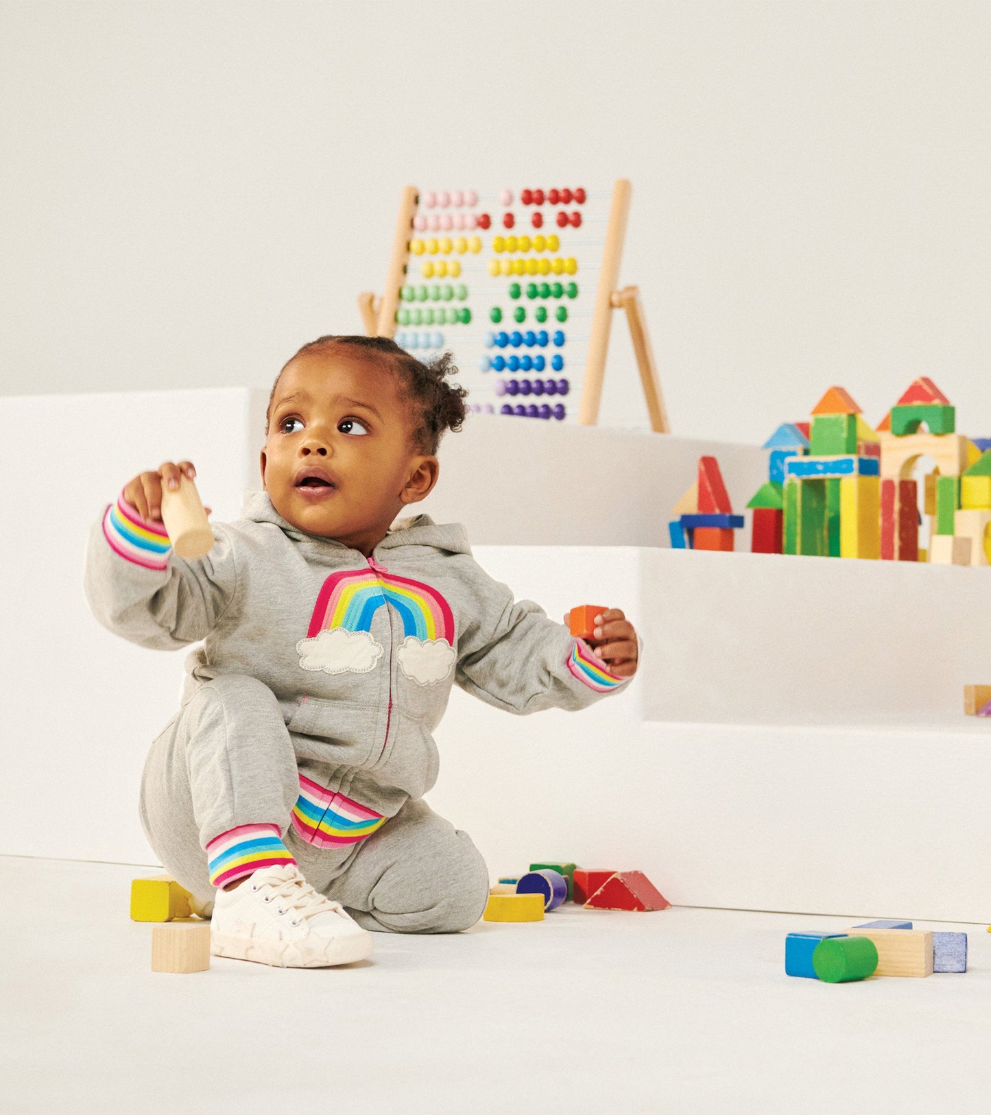 Gray full zip hoodie with rainbow and cloud design, featuring colorful striped cuffs, worn by a child playing with blocks.