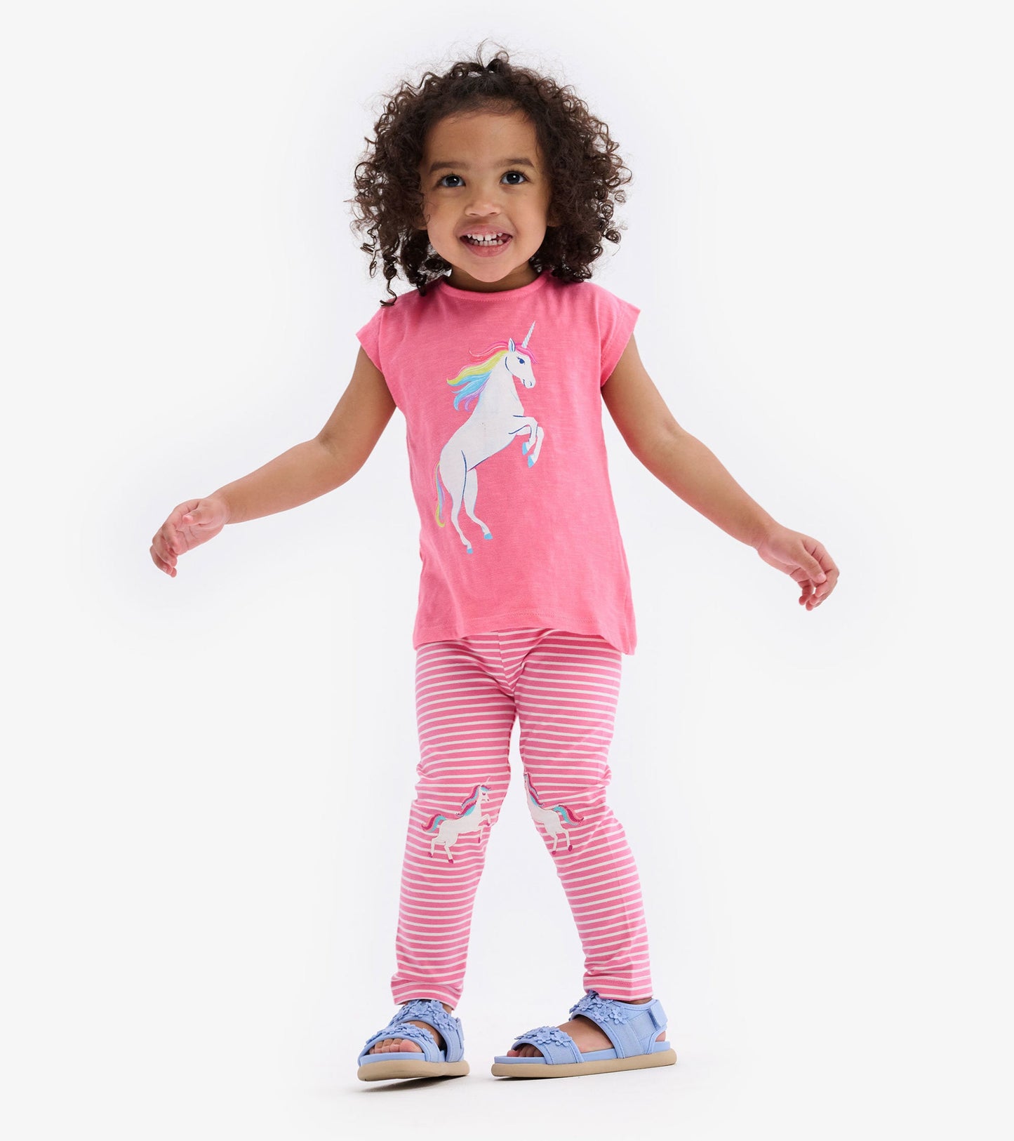 Pink toddler tee with unicorn graphic, paired with striped leggings featuring unicorns, worn with blue sandals.