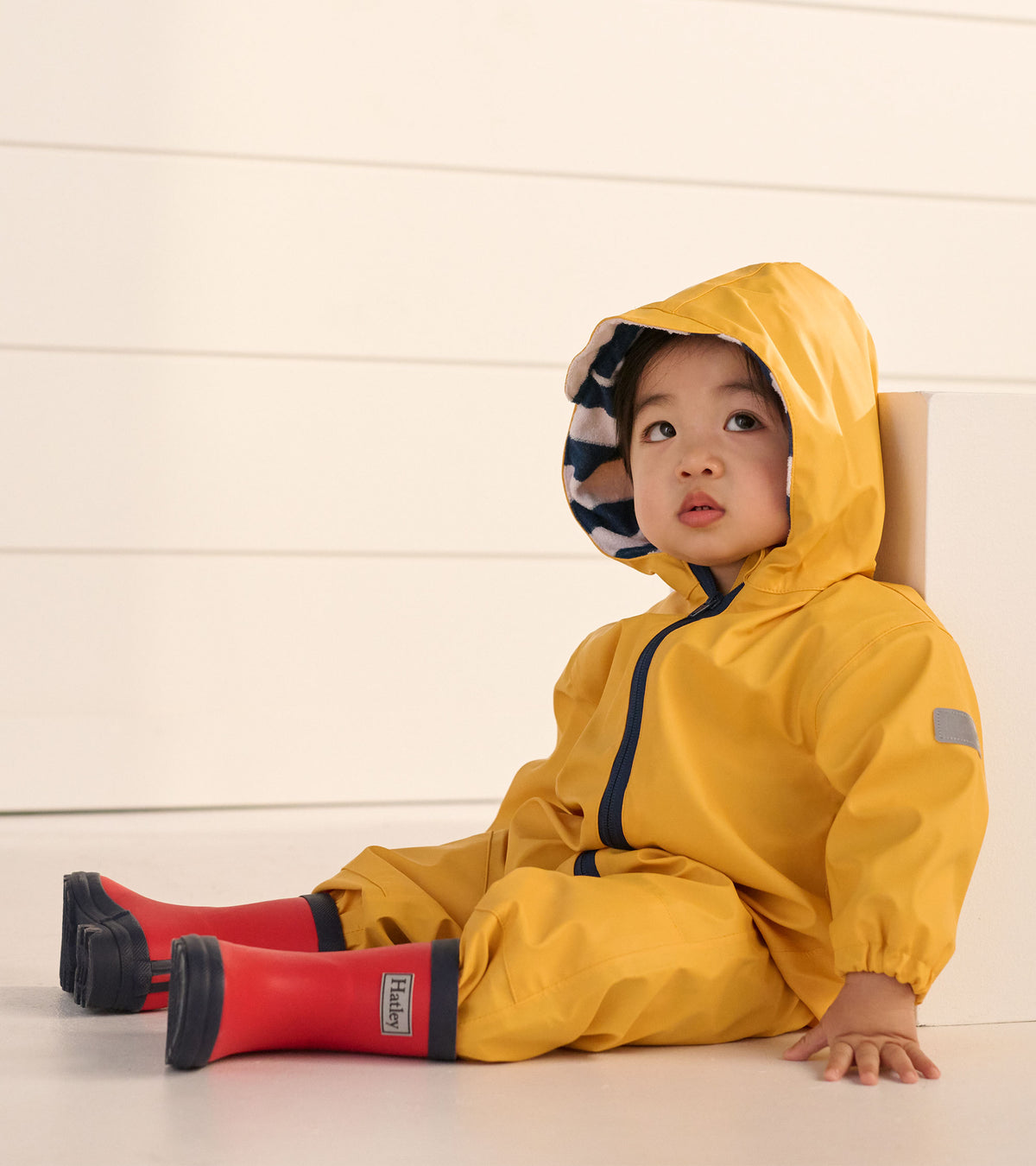 Toddler wearing a yellow hooded rain suit with striped hood lining and red rain boots, seated indoors