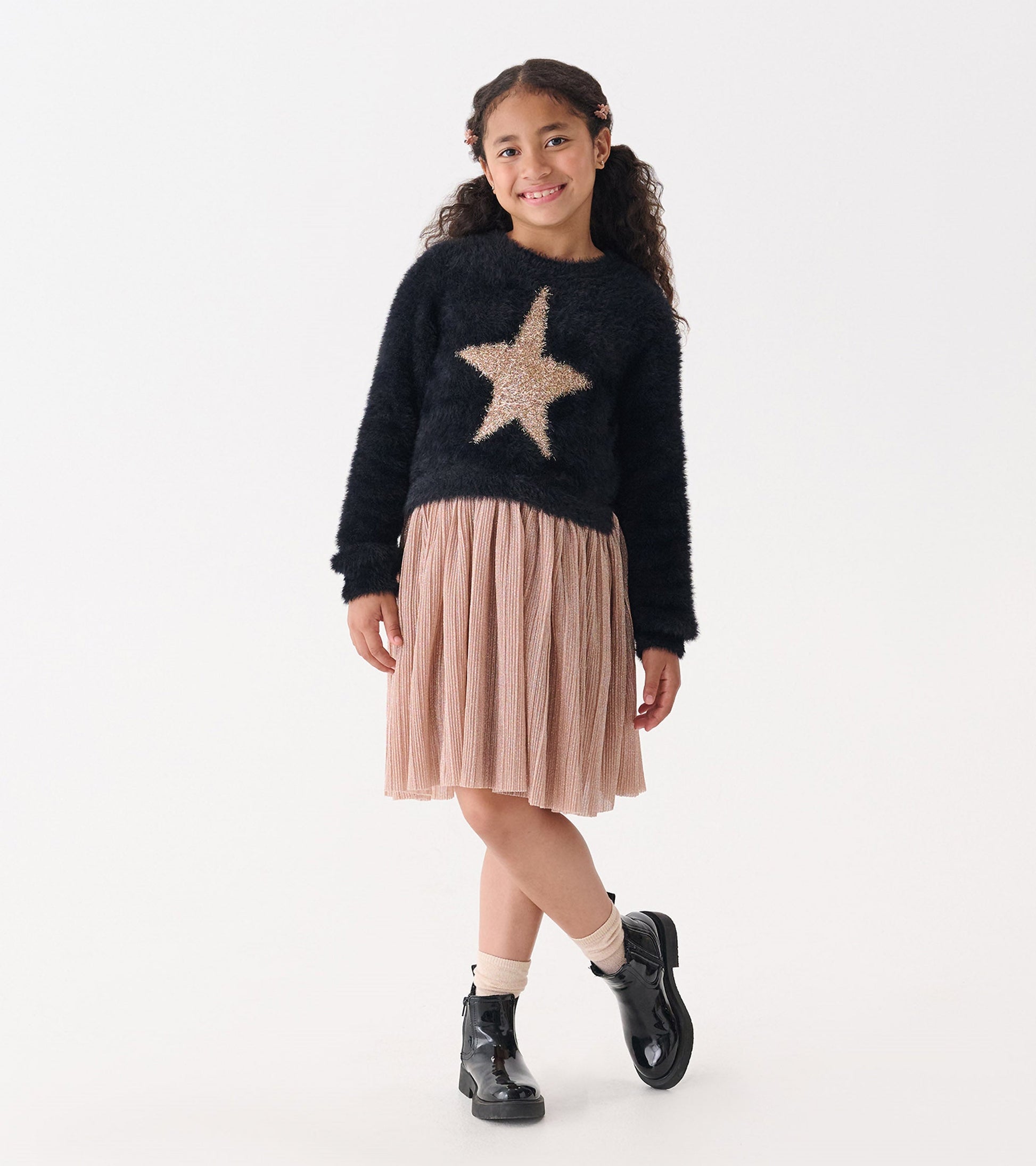 Black fuzzy sweater with gold star design paired with a pleated pink skirt and black ankle boots.