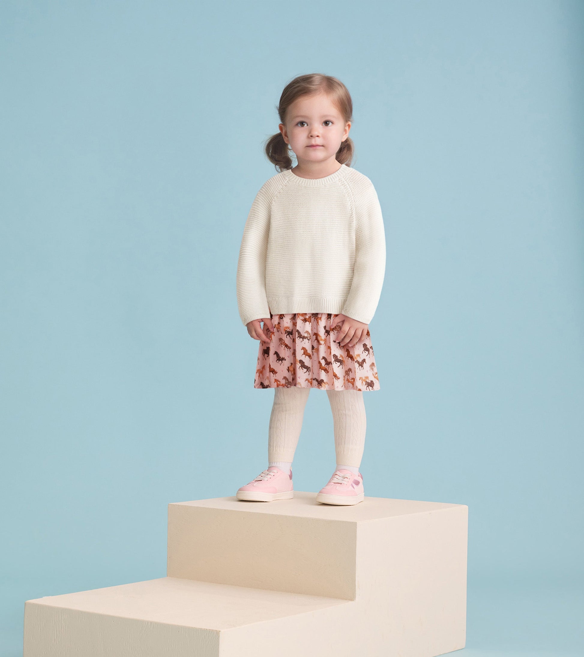 Cream sweater with a pink horse-patterned skirt, worn by a girl standing on a step, paired with light pink shoes.