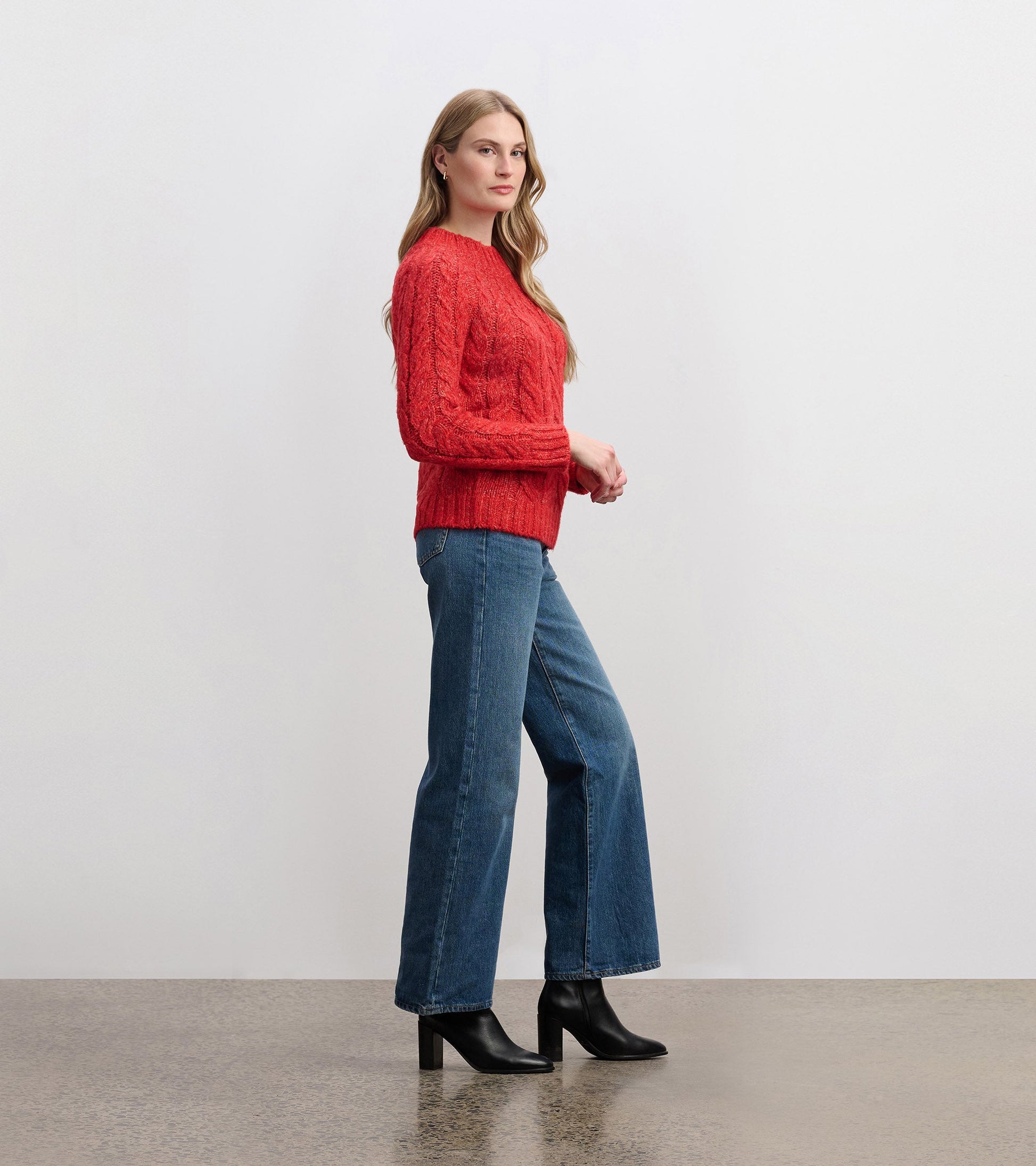Red cable knit jumper with long sleeves, styled with blue jeans and black heeled boots, shown from the side.