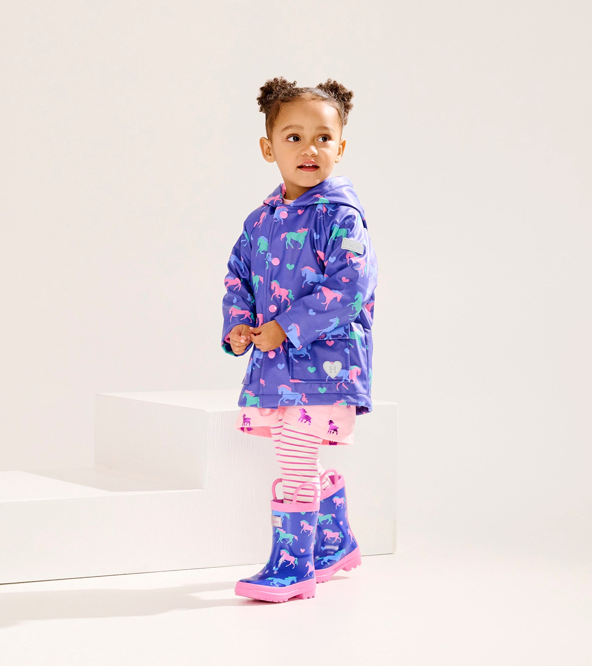 Purple button-up raincoat with horse and heart patterns, paired with pink striped leggings and matching rain boots.