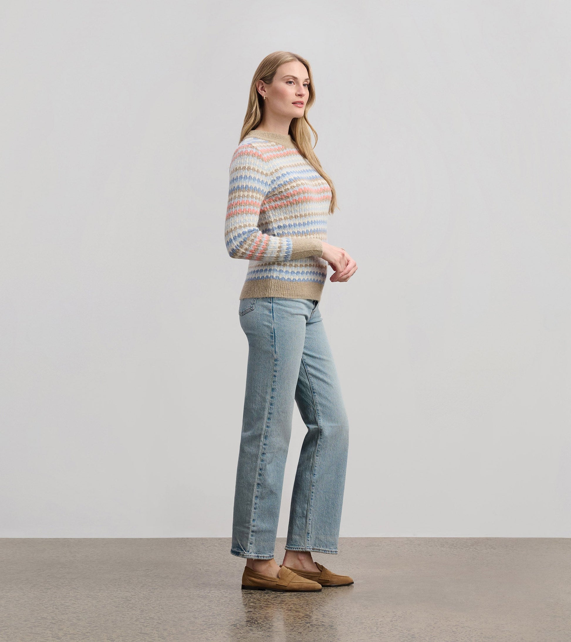 Multicolored striped jumper with a round neckline, paired with light blue jeans and brown slip-on shoes, shown from the side.