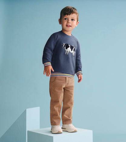 Navy pullover sweatshirt with a cow graphic and striped cuffs, worn by a boy standing on a blue platform.