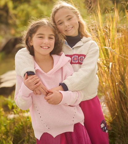 Girl wearing a light pink hooded pullover sweater with cream heart sleeves outdoors
