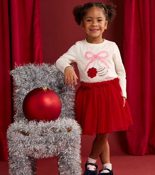Toddler girl wearing an ivory long-sleeve tee with pink bow and ornament graphic, styled with a red skirt