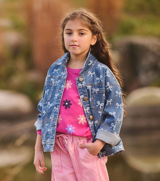 Girl wearing a blue slouchy denim jacket with allover white bow print and brass-tone buttons