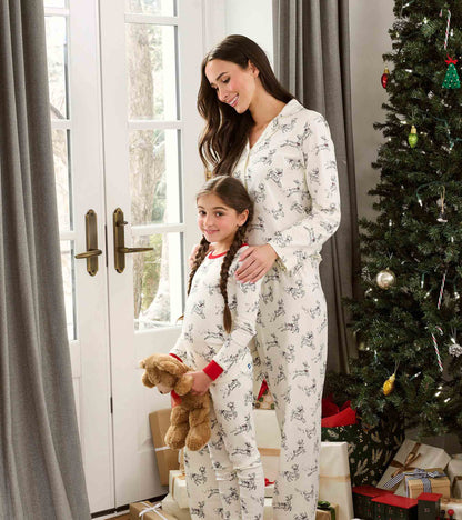 Girl wearing cream reindeer print pajamas with red trim, standing by a Christmas tree with an adult in matching print
