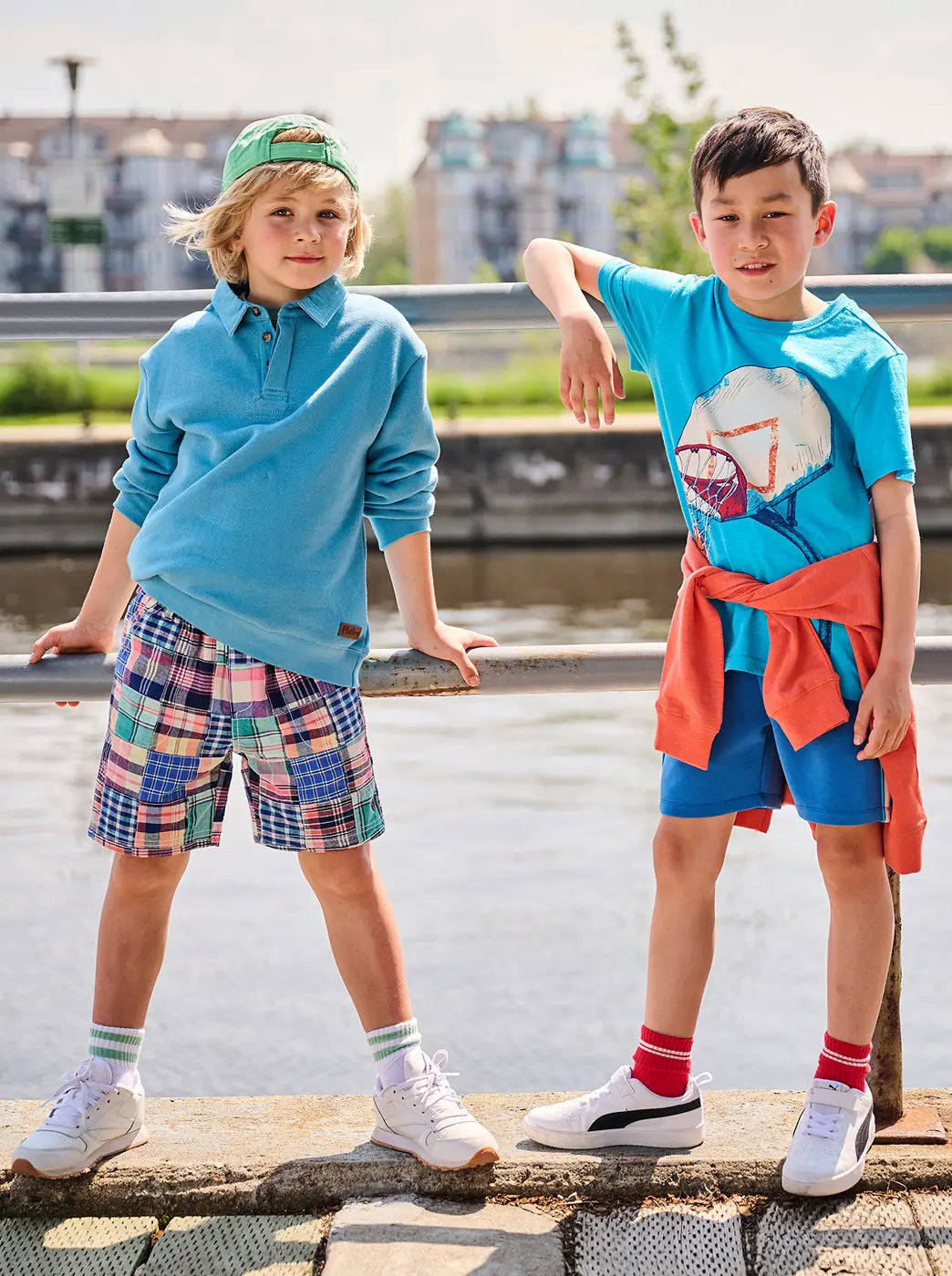 Promotes the boys collection. Showing two boys modeling bright casual outfits by a riverside railing.