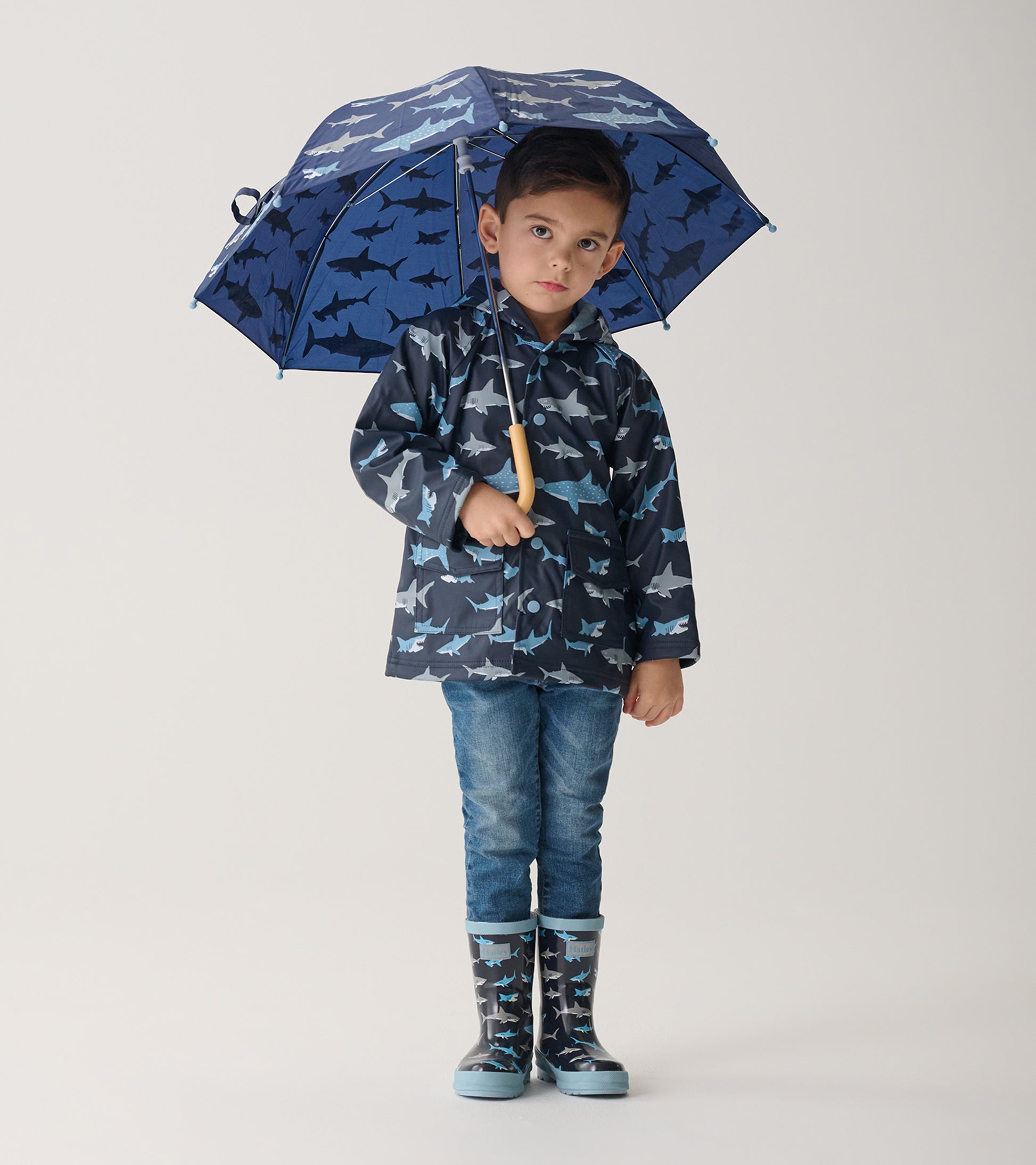 Child wearing navy shark print raincoat with matching rain boots, holding an open blue shark umbrella