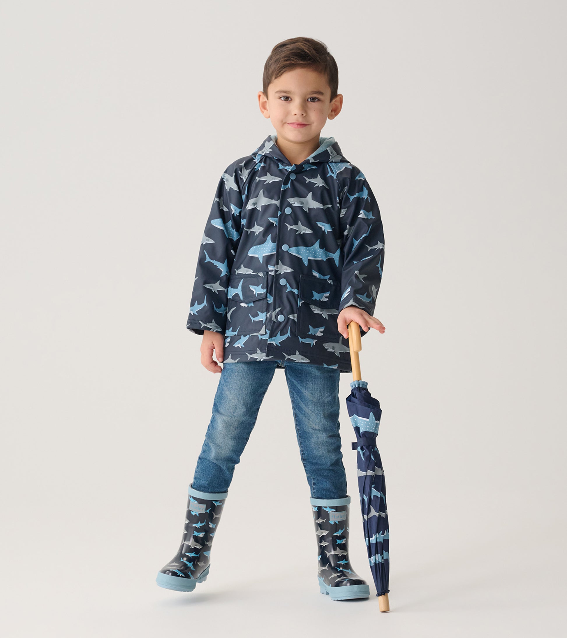 Child modeling navy shark print raincoat with matching rain boots, holding a closed umbrella