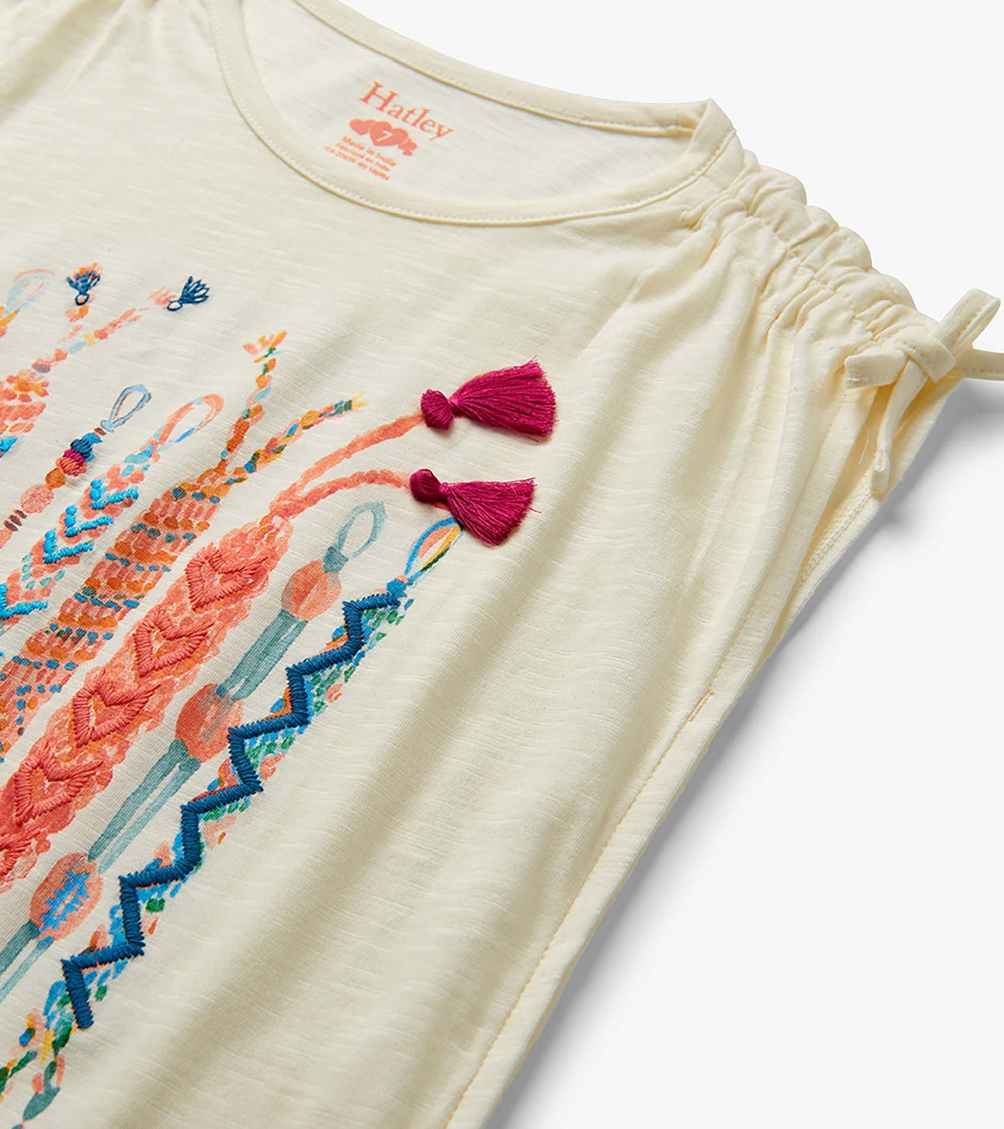 Cream girls top with colorful friendship bracelet print, cinched shoulder ties, and pink tassel details