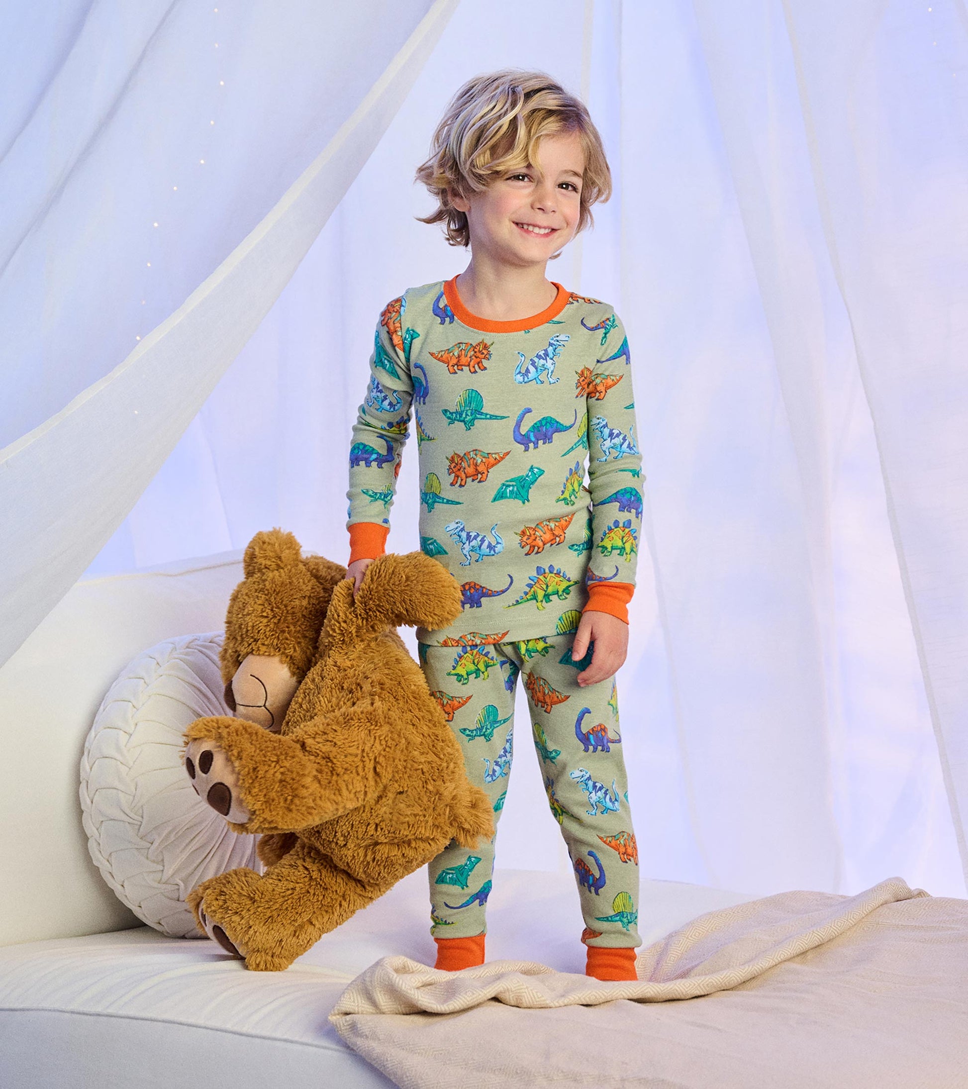 Child wearing sage green dinosaur print pajamas with bright orange trim, standing on a bed with a teddy bear