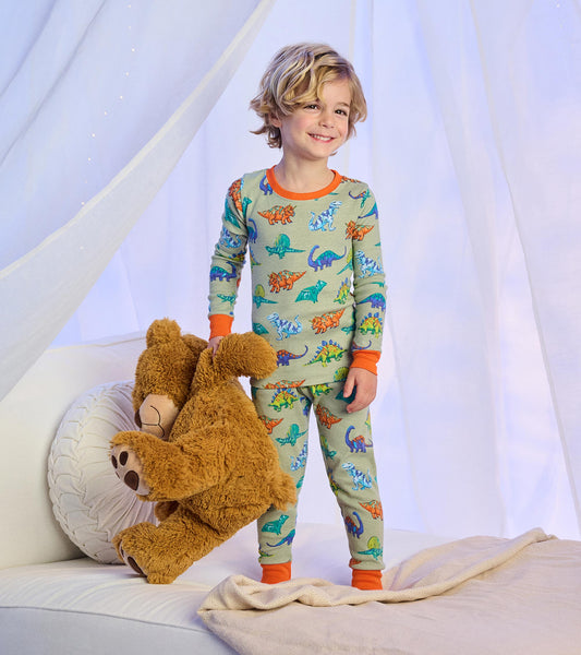 Child wearing sage green dinosaur print pajamas with bright orange trim, standing on a bed with a teddy bear