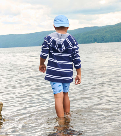 Back view of a boy wearing a navy and white striped hooded long-sleeve cover-up at the beach, with light blue shorts and cap