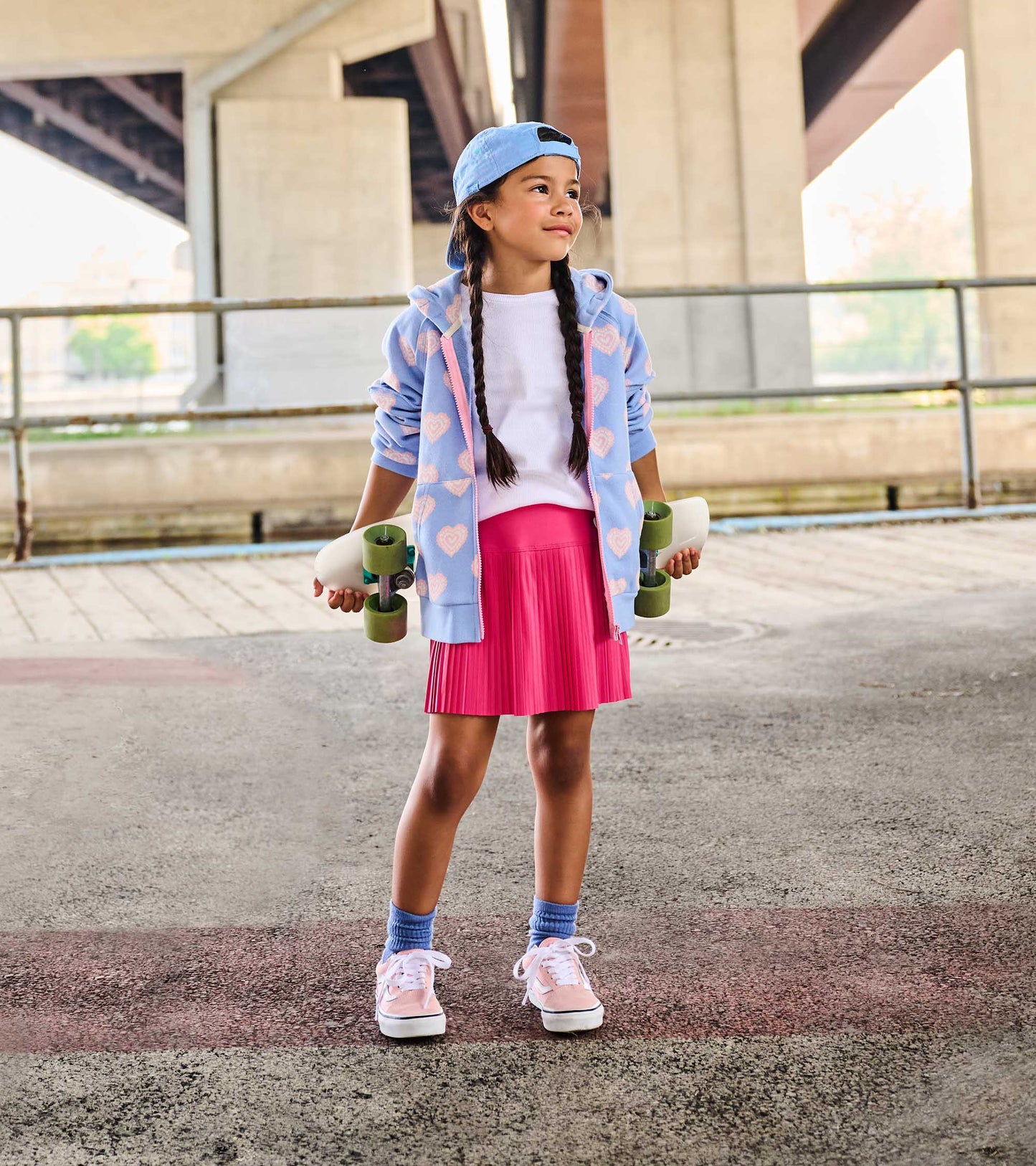 Girl wearing a bright pink pleated sporty skort with a white top and blue hoodie, holding a skateboard outdoors