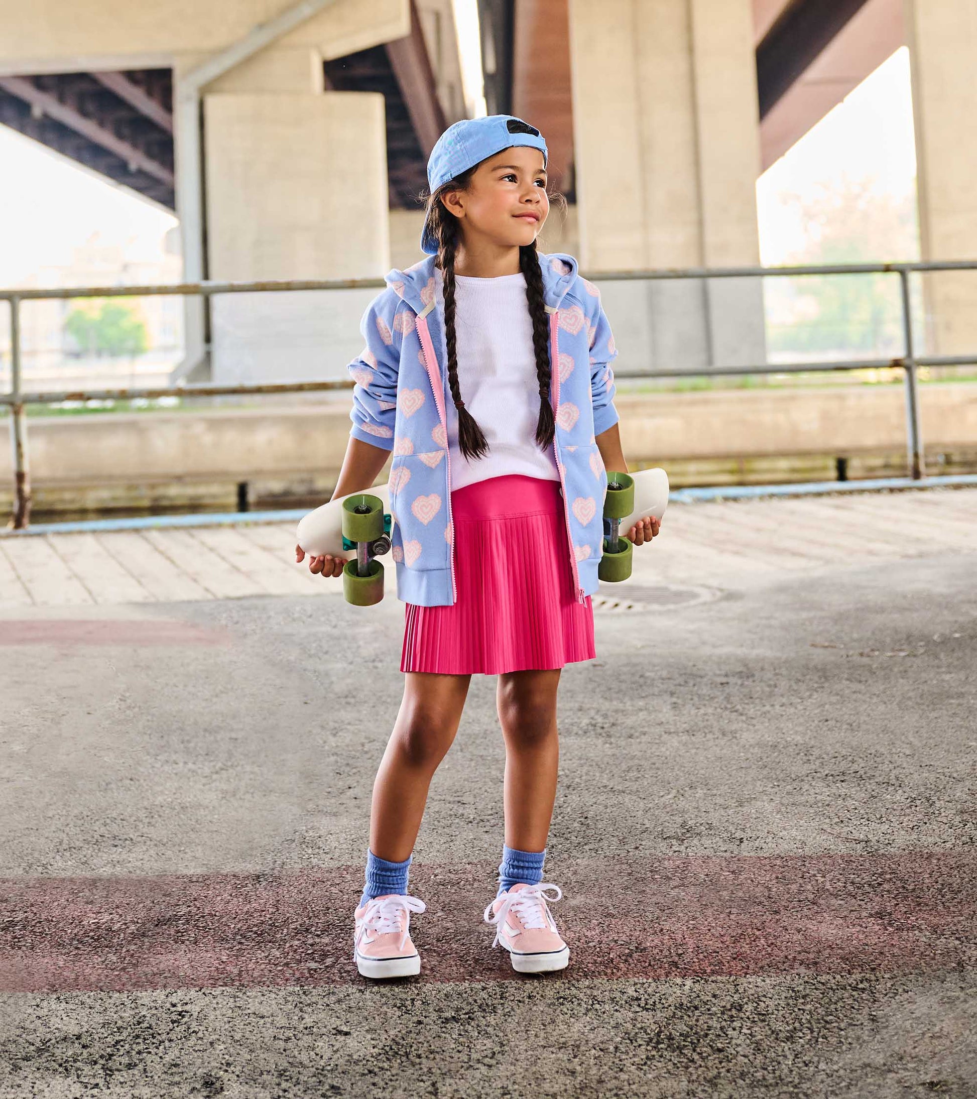 Girl wearing a bright pink pleated sporty skort with a white top and blue hoodie, holding a skateboard outdoors