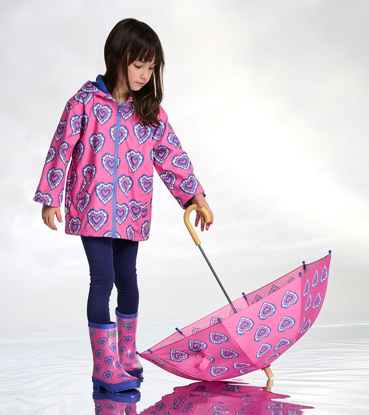 Girl wearing a pink zip-up raincoat with blue heart print, shown with matching umbrella and rain boots.