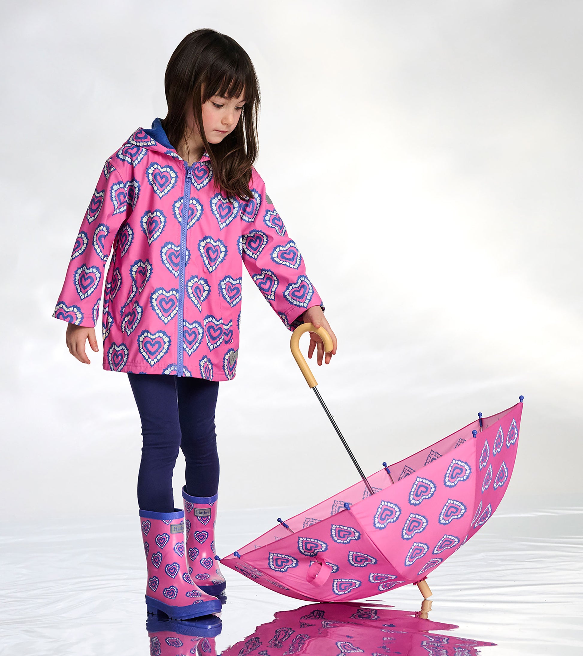 Girl wearing a pink zip-up raincoat with blue heart print, shown with matching umbrella and rain boots.