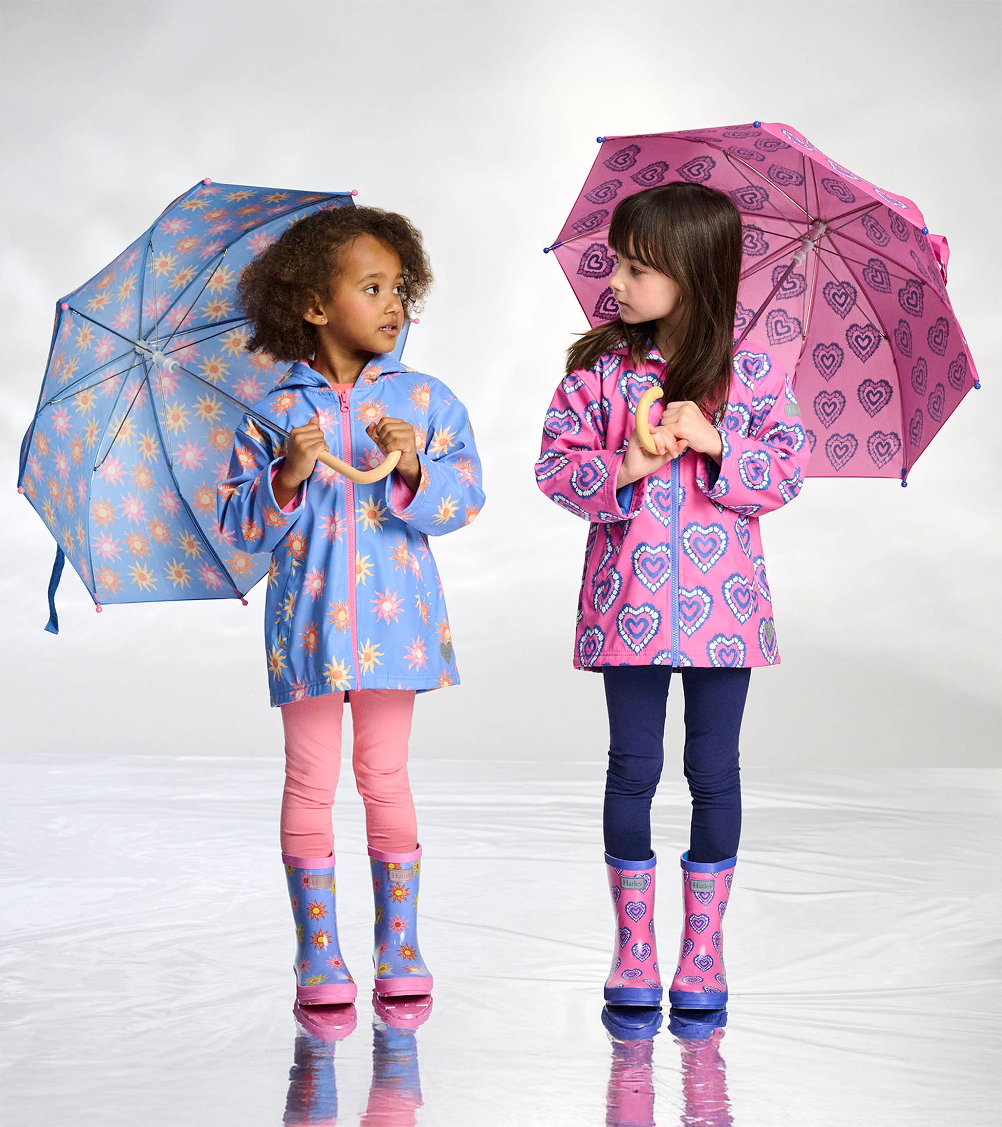 Girls zip-up raincoats with color-changing heart and sunburst prints, shown on two children holding umbrellas