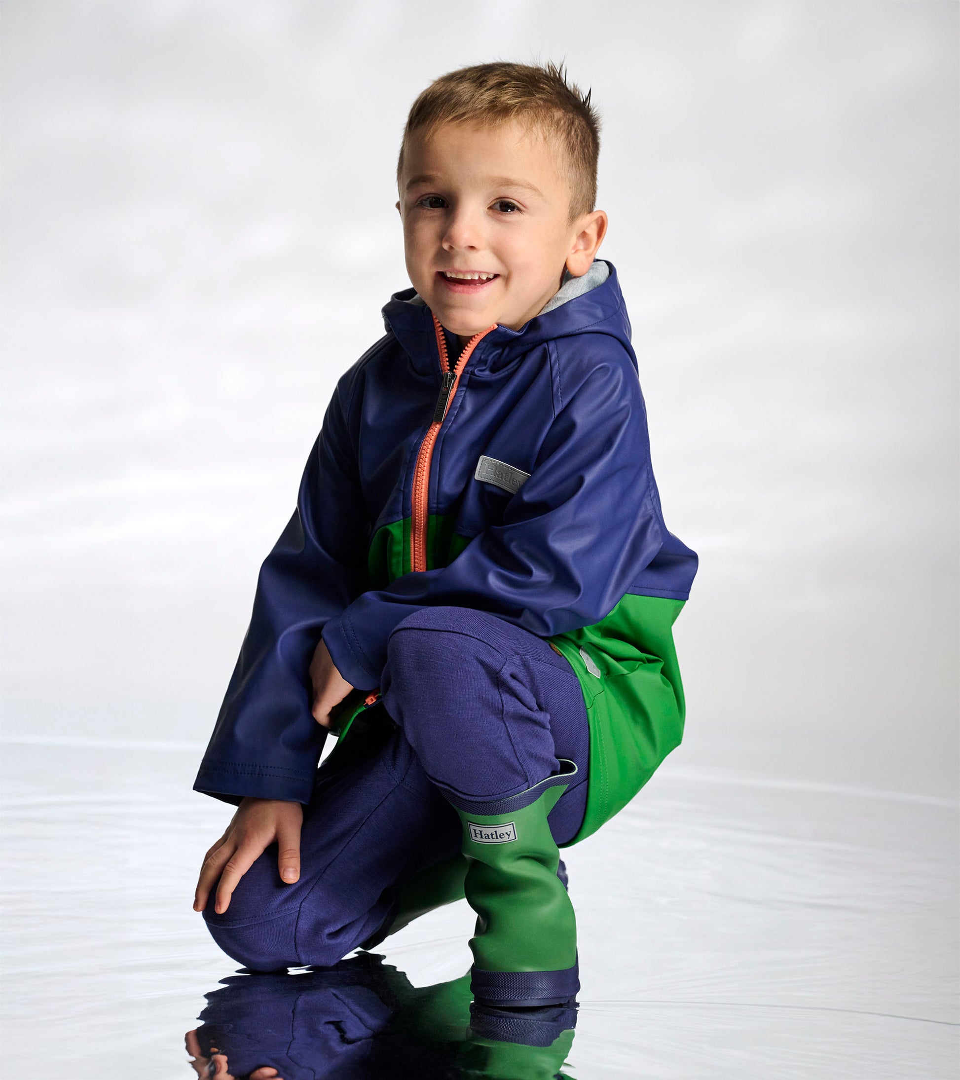 Boy wearing a navy and green colorblock hooded raincoat with orange zipper, kneeling front view