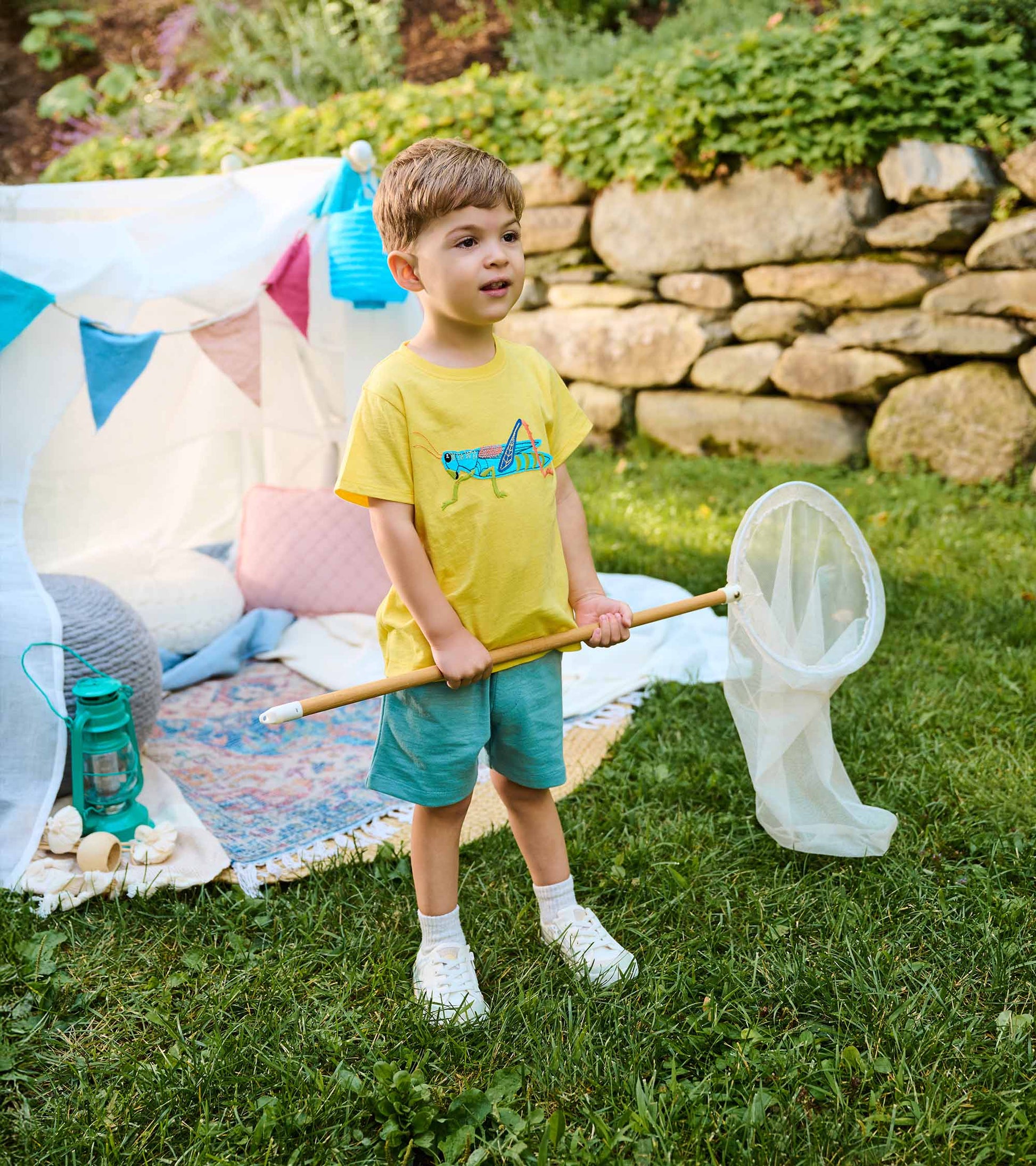 Toddler wearing a yellow grasshopper T-shirt with light blue shorts, standing outdoors holding a bug net