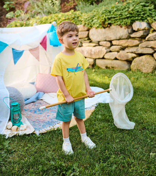 Toddler wearing a yellow grasshopper T-shirt with light blue shorts, standing outdoors holding a bug net