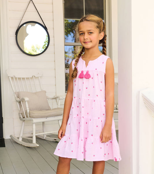 Girl wearing a pink gingham tiered dress with small berry print, sleeveless cut, and tassel ties