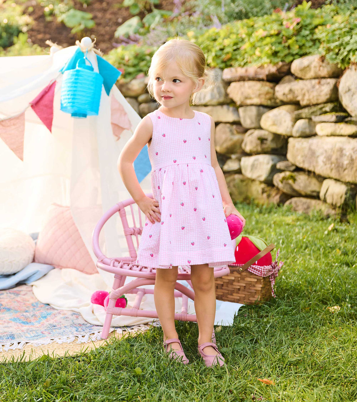 Toddler modeling pink and white gingham strawberry dress beside a pink chair outdoors