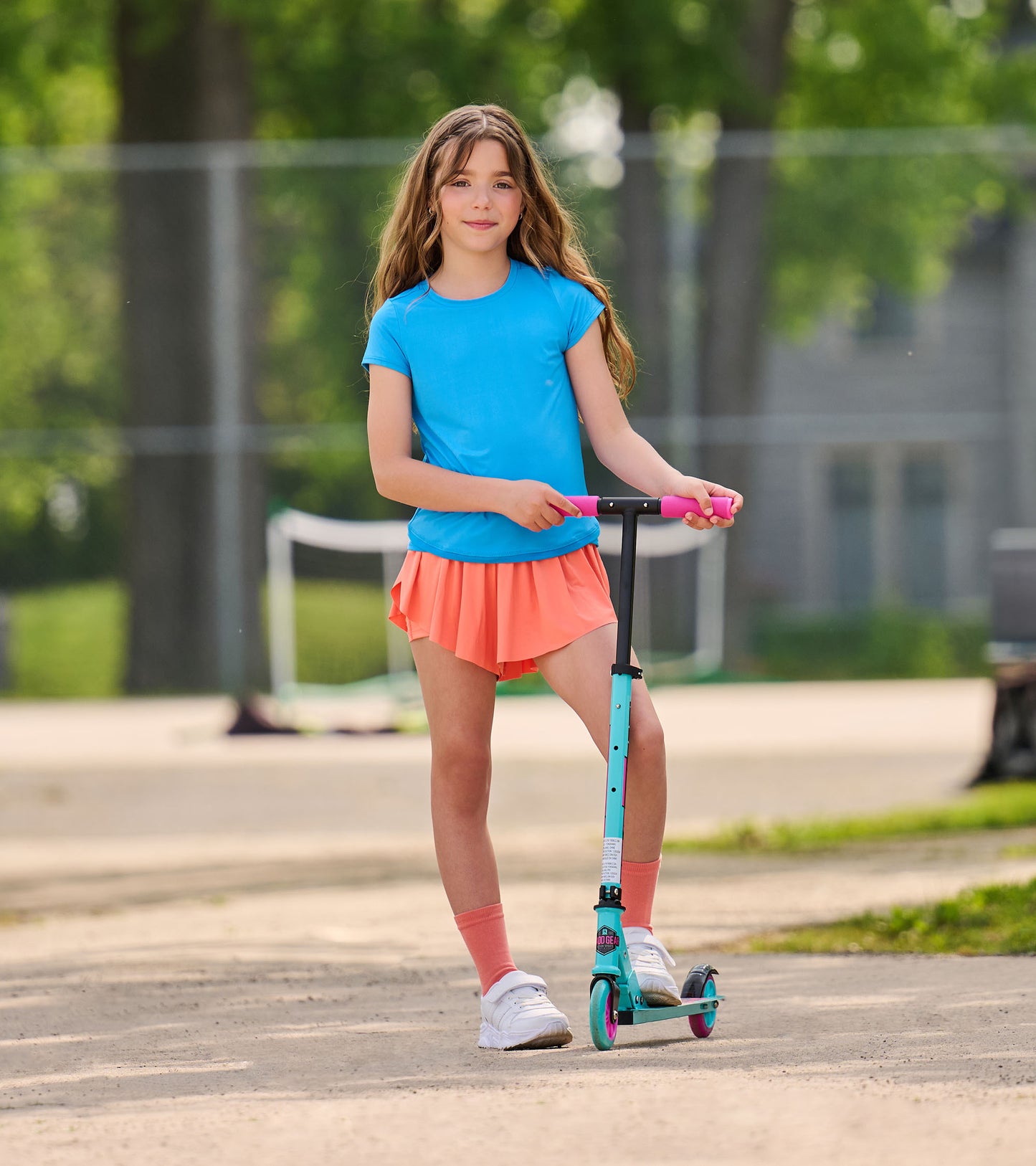 Girl wearing a bright blue short-sleeve T-shirt with coral shorts, standing on a scooter outdoors.