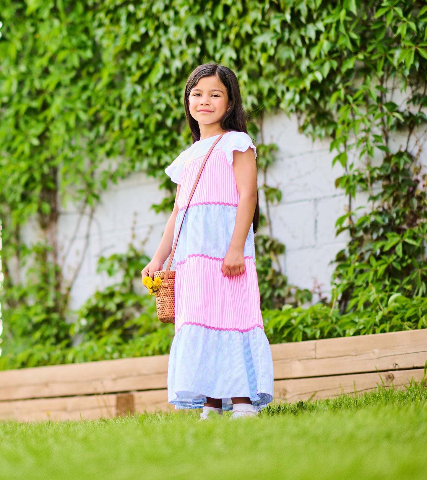 Side view of girl wearing a pink and blue tiered striped midi dress with pink trim outdoors