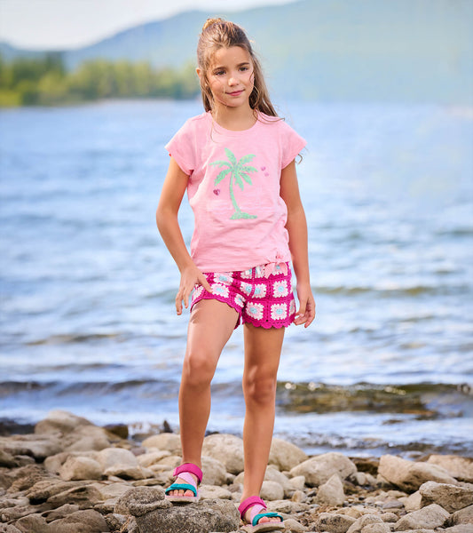 Girl wearing a pink short sleeve tie-front T-shirt with palm tree graphic, shown outdoors by the water